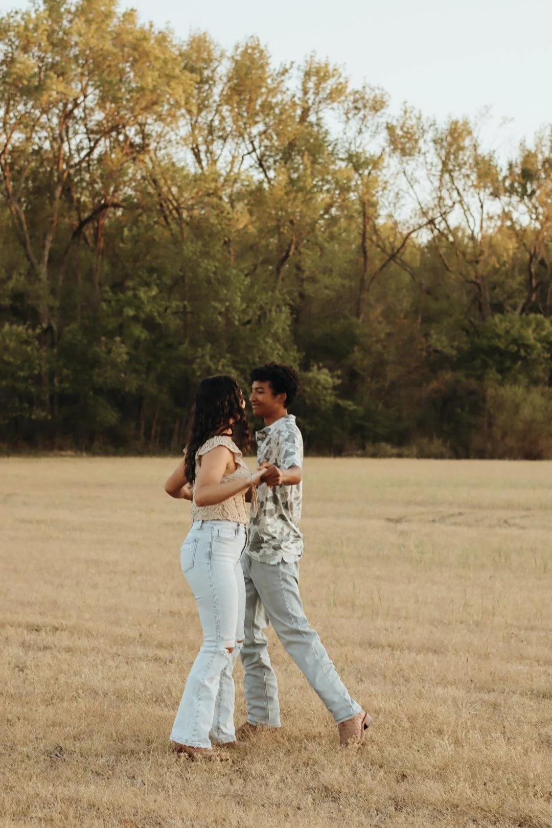 A young couple dancing and holding hands in a grassy field with a background of trees and a clear sky at sunset.