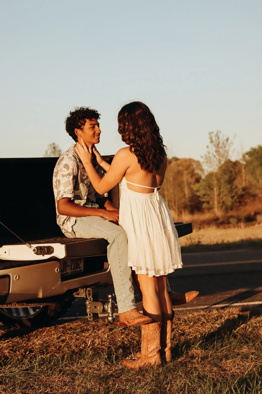 A woman and a man sharing an intimate moment near a vintage truck in an outdoor setting. The woman is standing, wearing a white dress and cowboy boots, while the man is sitting on the truck's tailgate, dressed casually in a patterned shirt and jeans.