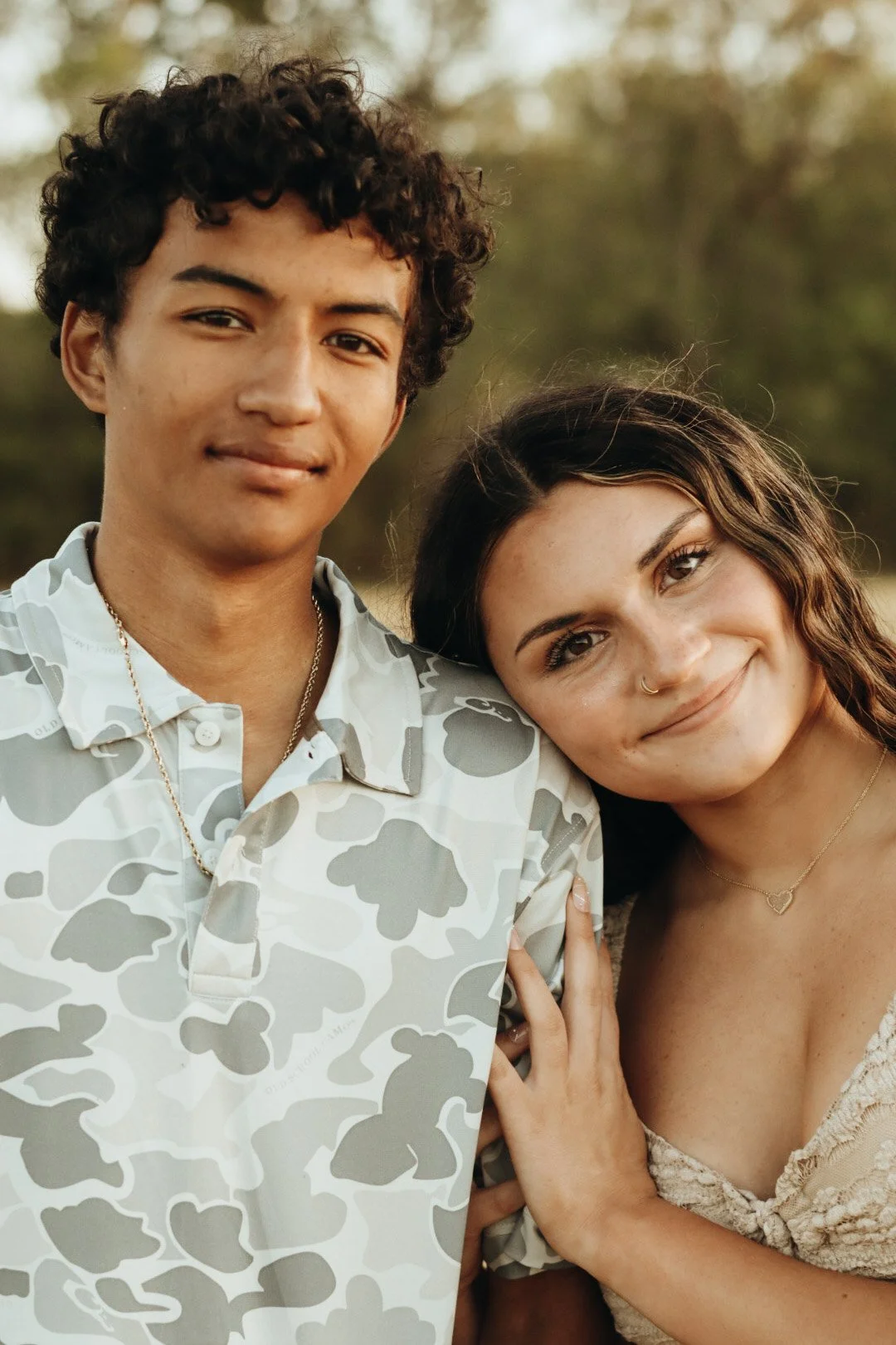 A young man with curly hair and a young woman with wavy hair, both smiling, posing outdoors during what appears to be sunset, with blurred trees in the background.