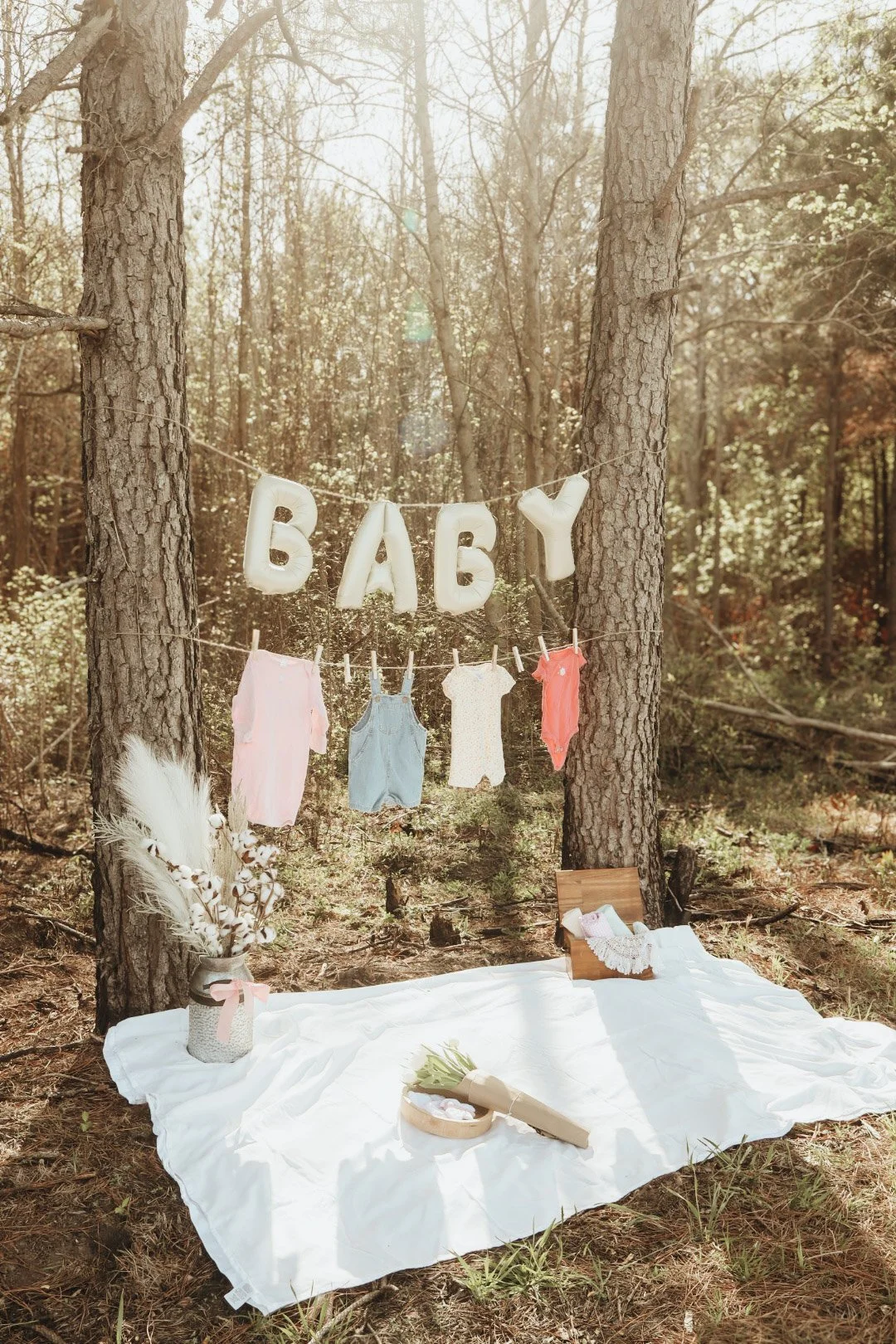 Outdoor baby shower setup in the woods with a white blanket, hanging baby clothes, and a decorated banner spelling 'BABY'.