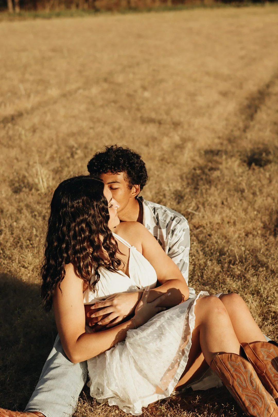 A young couple sitting on a grassy field, embracing, with the man gently kissing the woman's forehead, during sunset.