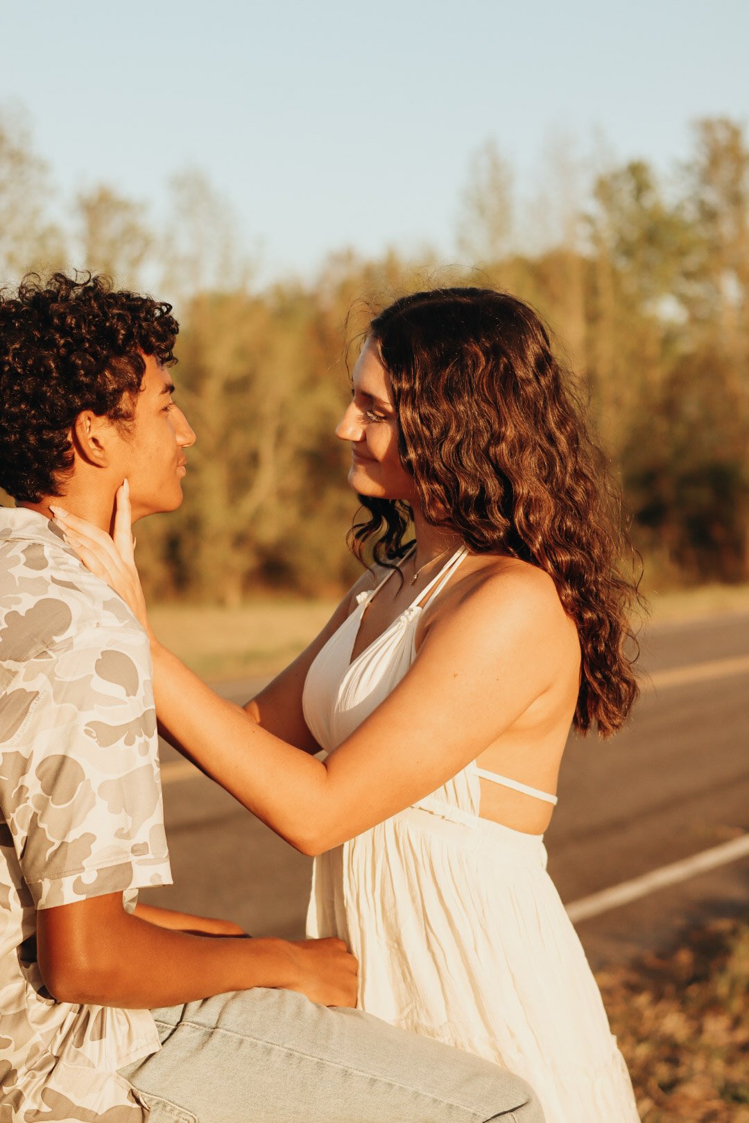 A man and woman face to face outdoors, with the woman gently holding the man's chin, both gazing into each other's eyes, during sunset.