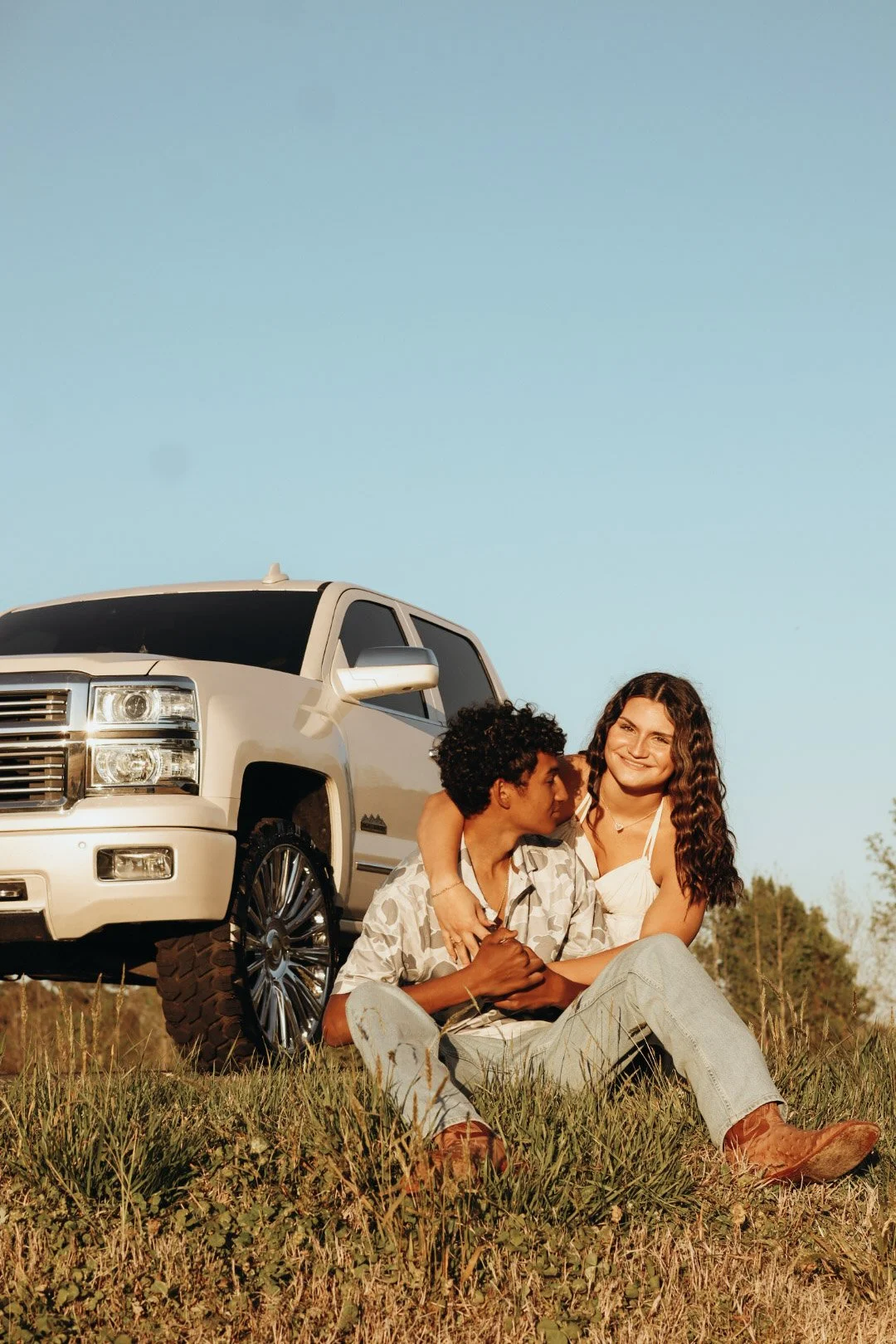 A happy couple sits on the grass next to a white SUV during a sunny day, embracing each other with trees in the background.
