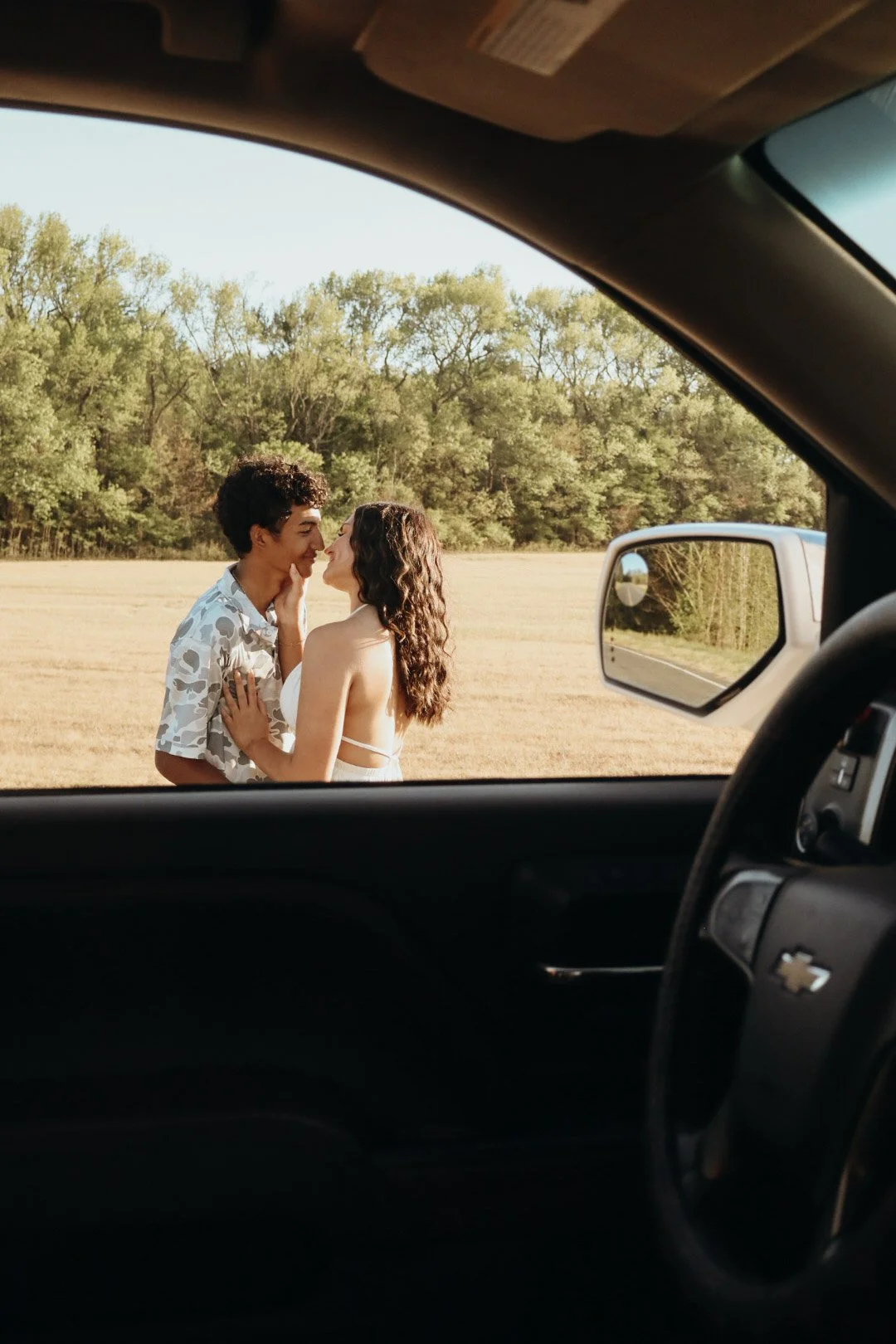 A young couple is standing outside of a vehicle, facing each other, with their noses touching and smiling. The scene is in a grassy field with trees in the background, taken through the window of a car.