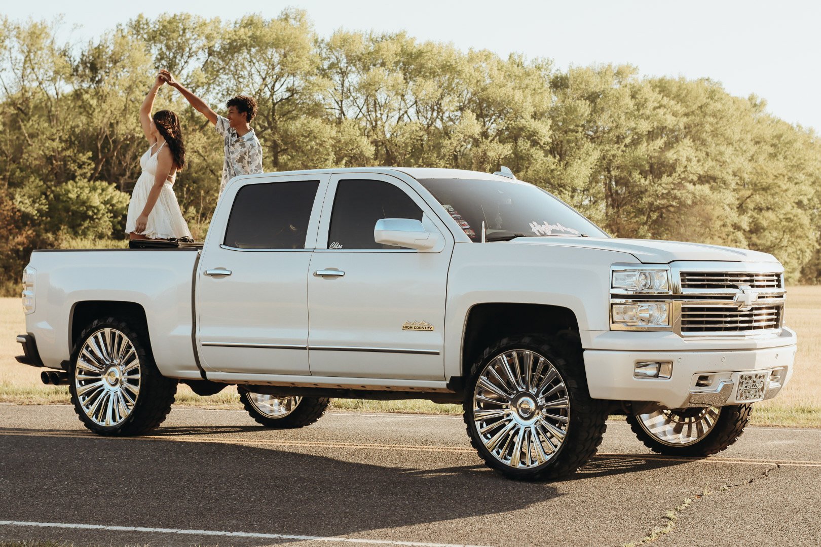 A white pickup truck with large, shiny, custom rims parked on the side of a rural road. Two people are standing in the truck bed, dancing and holding hands, with trees and a clear sky in the background.