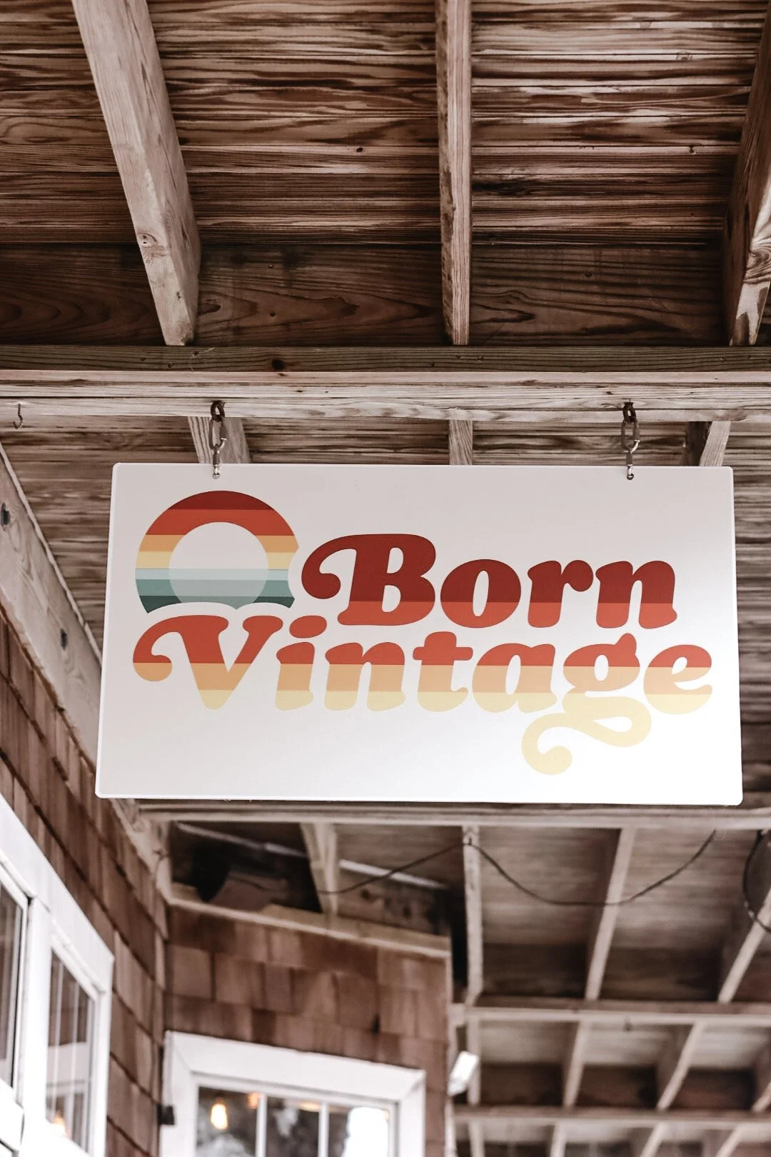 A white sign with colorful rainbow-like pattern and red text that says 'Born Vintage' hanging on a wooden outdoor ceiling.