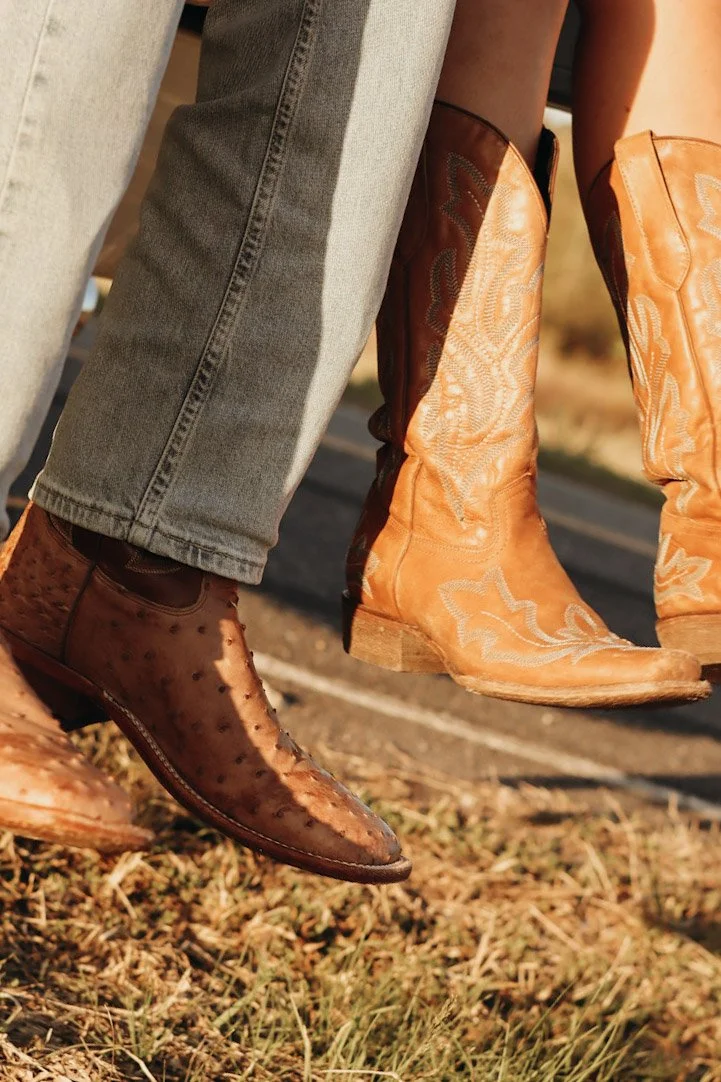 Close-up of people sitting on a fence, wearing cowboy boots and jeans, with dried grass on the ground and a road in the background.