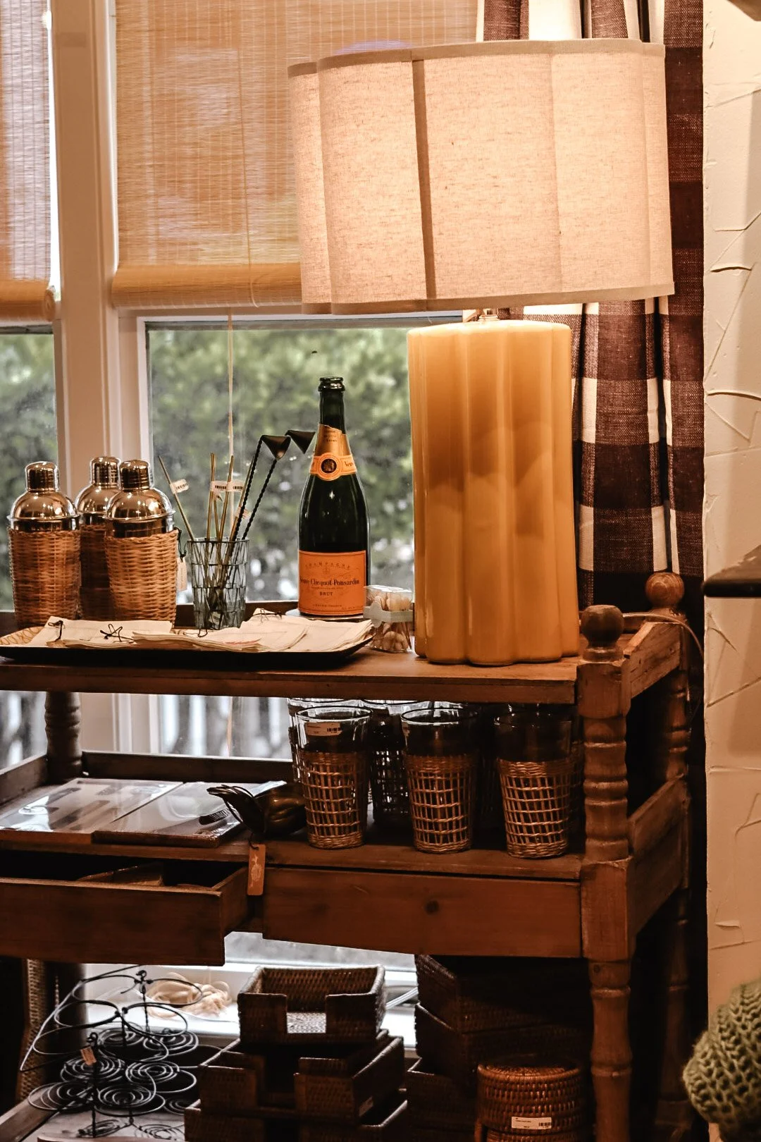A wooden side table by a window holds three metal bottles, a bunch of cocktail stirrers, and a bottle of sparkling wine. On the lower shelf, there are four wicker glasses and some outdoor-style tableware. Behind the table, there are bamboo blinds, a 