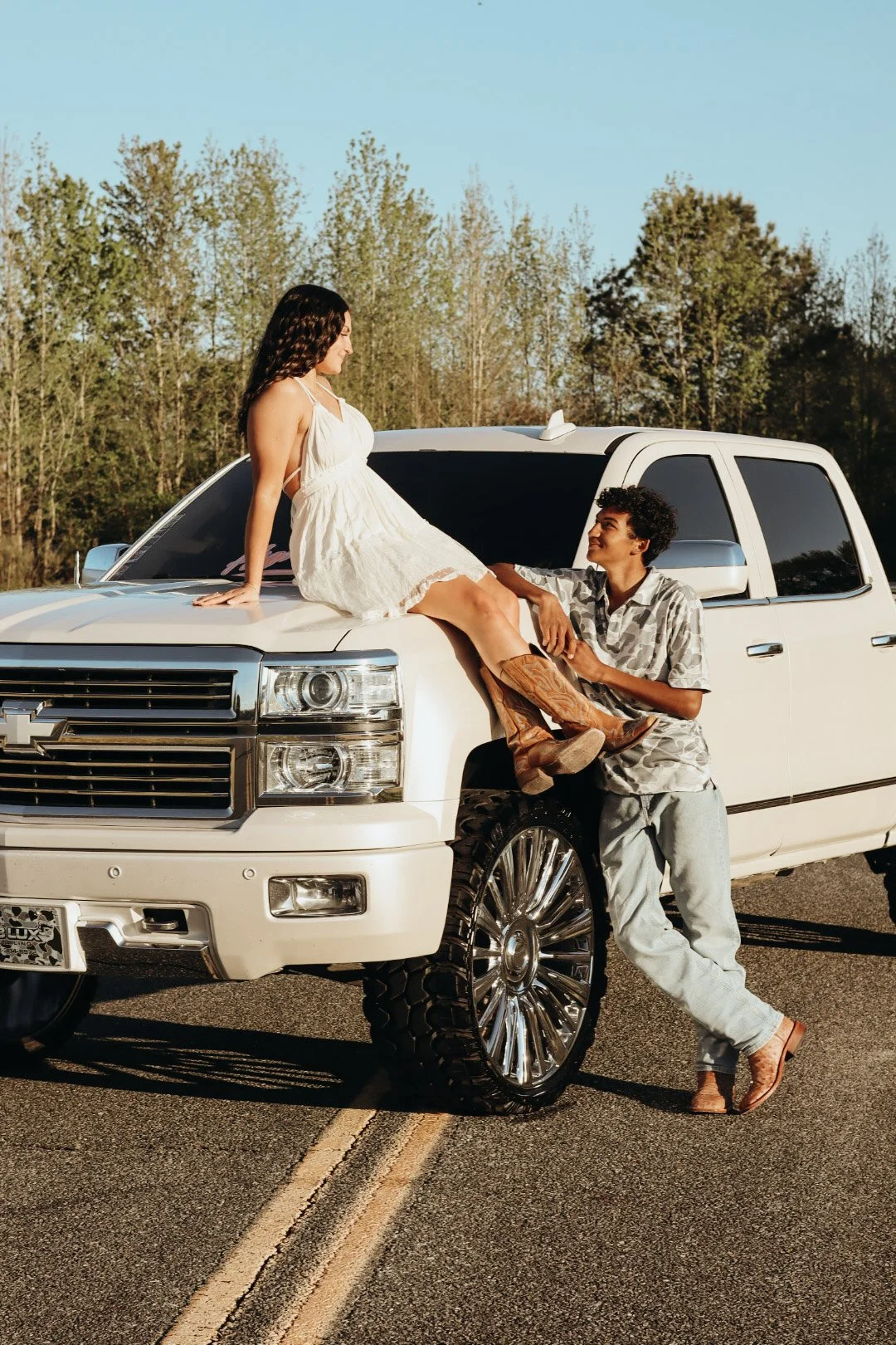 A young woman in a white dress and cowboy boots sitting on the hood of a white pickup truck, while a young man in a patterned shirt and light pants stands next to her and holds her hand, on an open road with trees in the background during golden hour