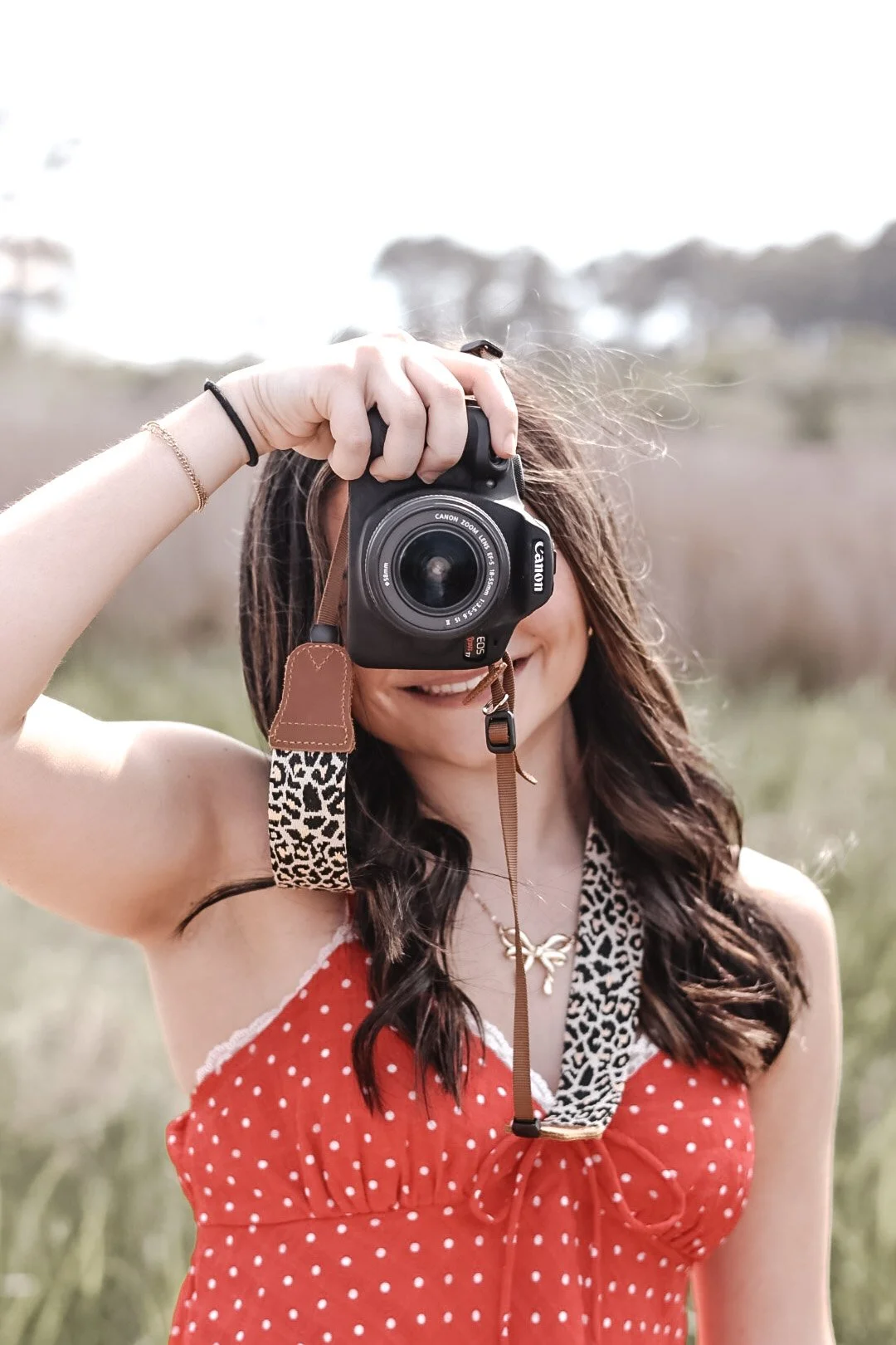 Woman holding a camera in front of her face, outdoors, smiling, wearing a red polka dot dress, with long wavy hair.
