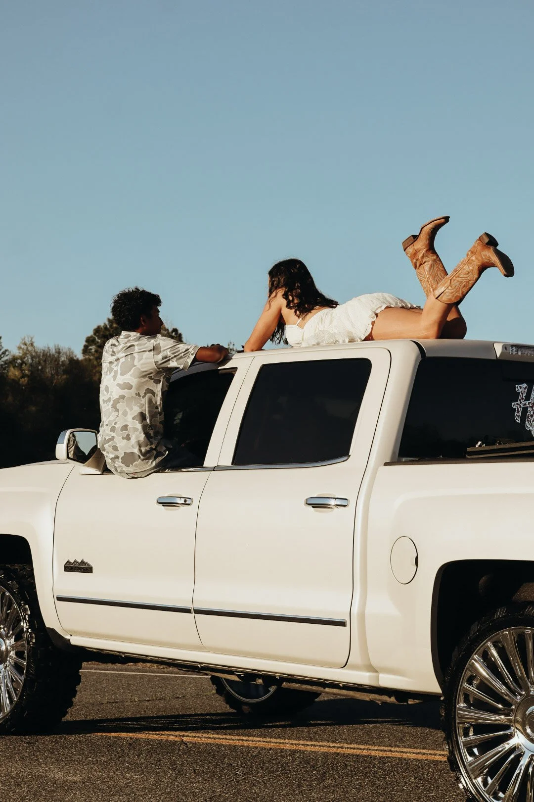 A young woman in a white dress and cowboy boots lying on the roof of a white SUV, talking to a young man sitting on the window edge. The SUV is parked outdoors under a clear blue sky.