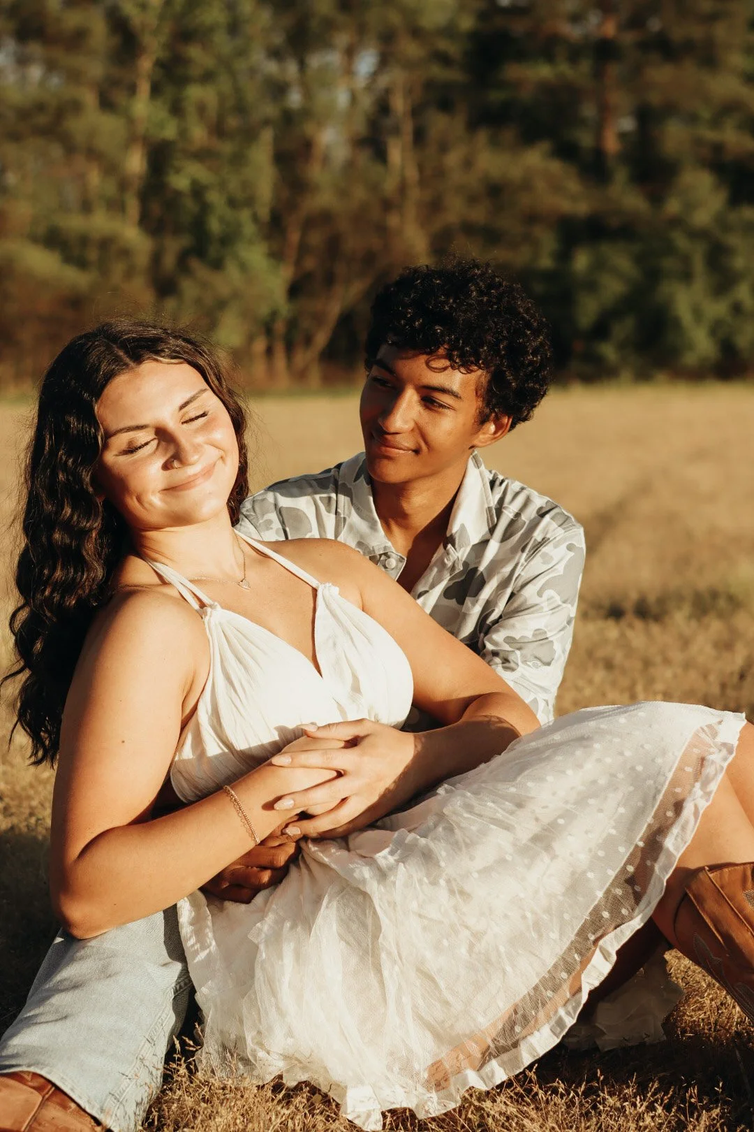 A happy couple enjoying a moment outdoors in a field with trees in the background. The woman, wearing a white dress, is smiling with her eyes closed as the man, in patterned shirt, looks at her affectionately while holding her in his arms.