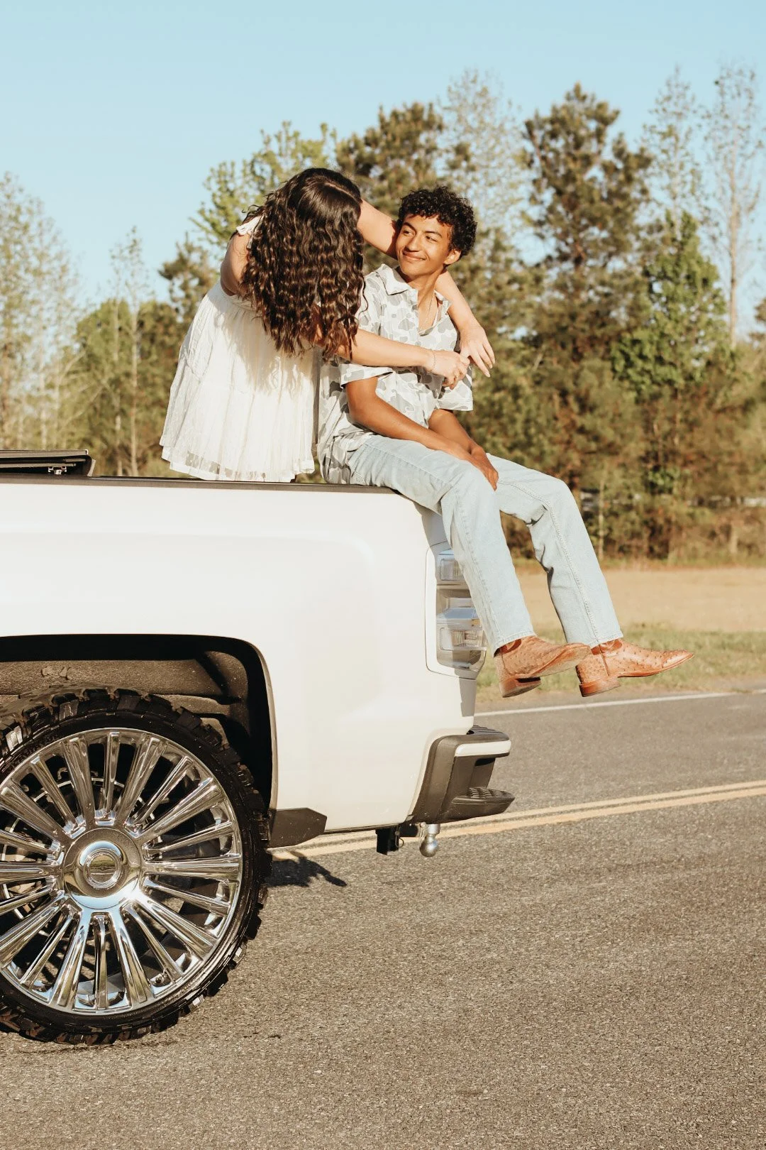 A young man and woman sitting on the edge of a white pickup truck's bed, with the woman leaning over to touch the man's face. They are outdoors on a sunny day, with trees and a clear sky in the background.