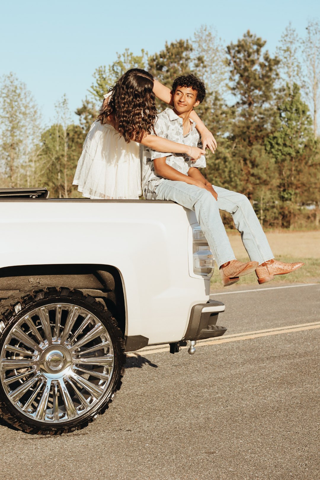 Two young women sitting on the back of a white pickup truck, outdoors on a rural road, with trees in the background. One woman with curly brown hair wears a white dress, and the other woman with short curly hair wears a patterned shirt and jeans. The