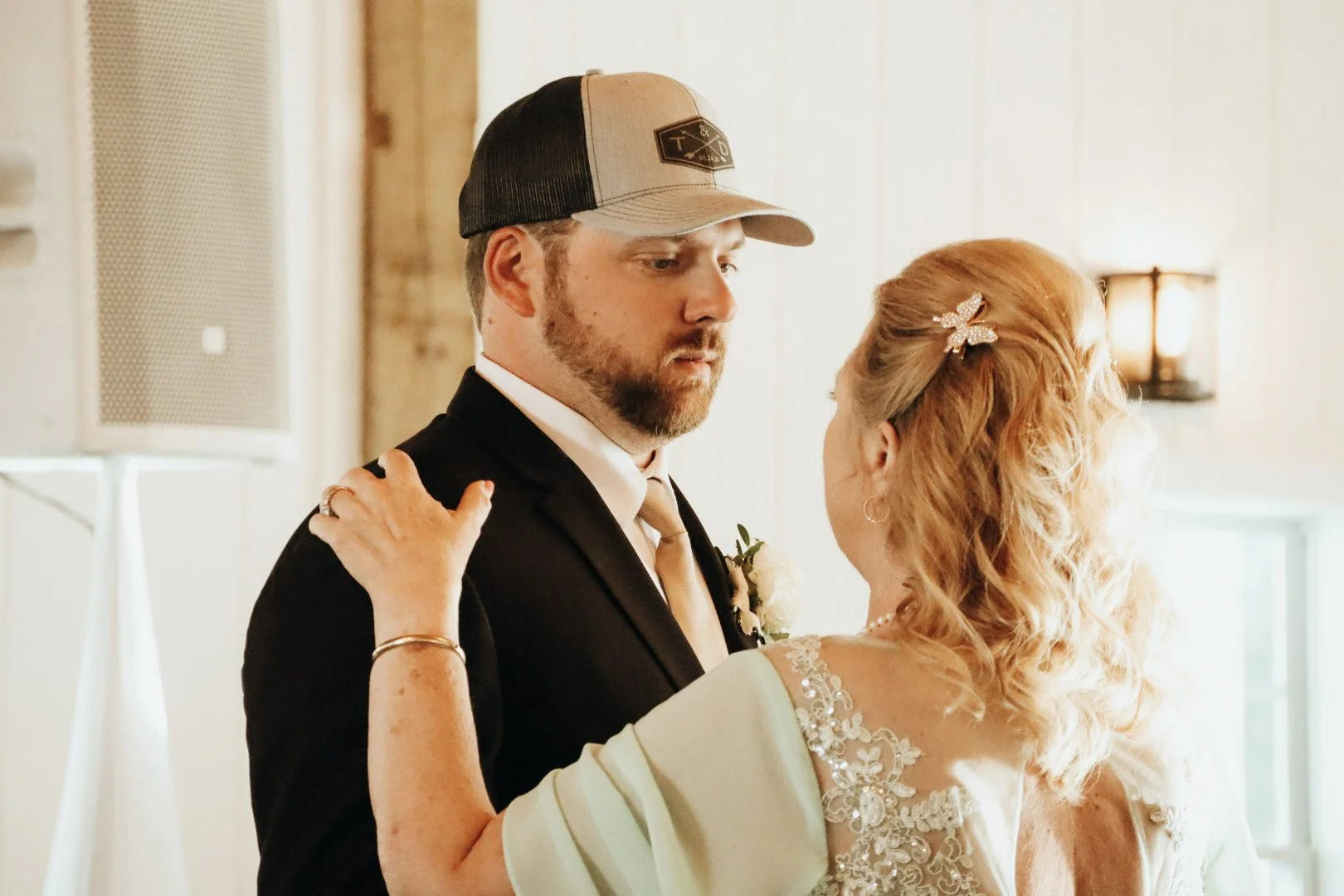 A man in a black tuxedo and a baseball cap dance with a woman in a light-colored dress with lace details, both appearing to share a special moment indoors with warm lighting.