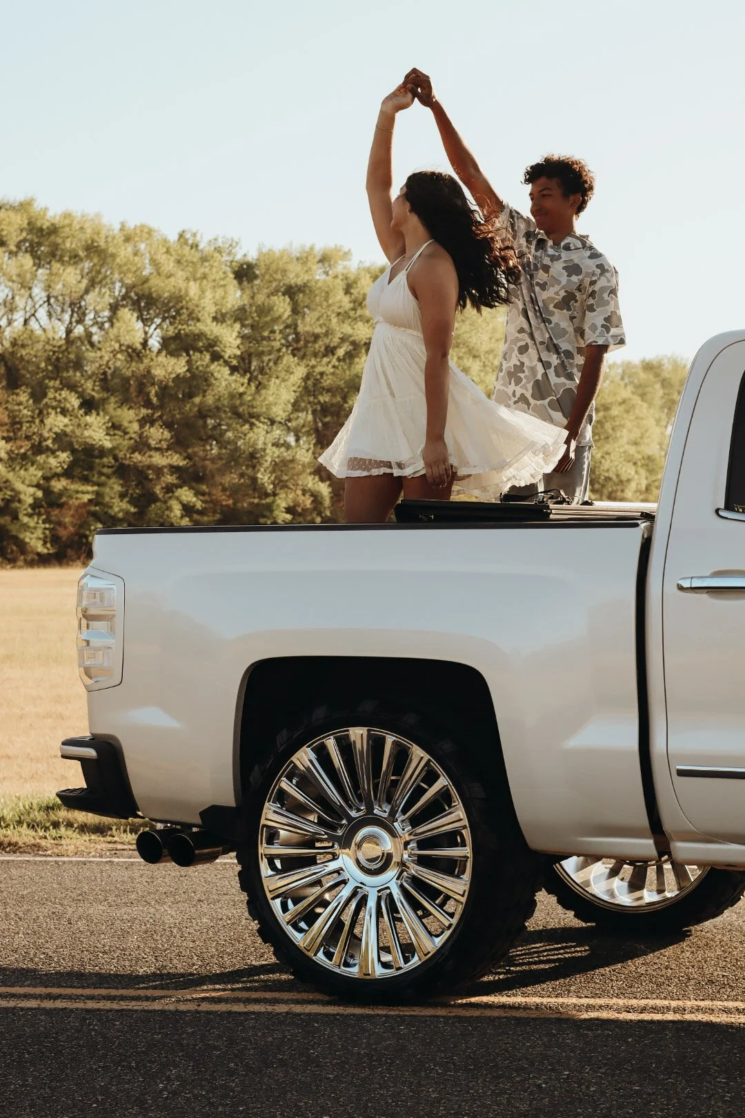 A woman and a man dancing in the back of a white pickup truck in an open field with trees in the background. The woman is wearing a white dress and the man a patterned shirt. The scene is set during sunset or late afternoon.