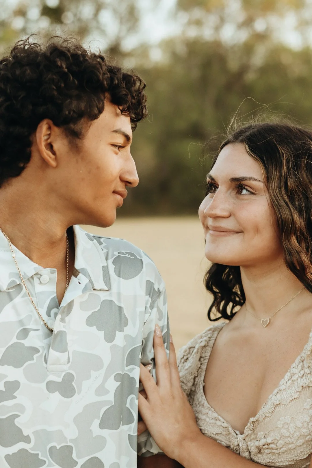 A young man and woman face each other outdoors, smiling softly, with trees in the background.