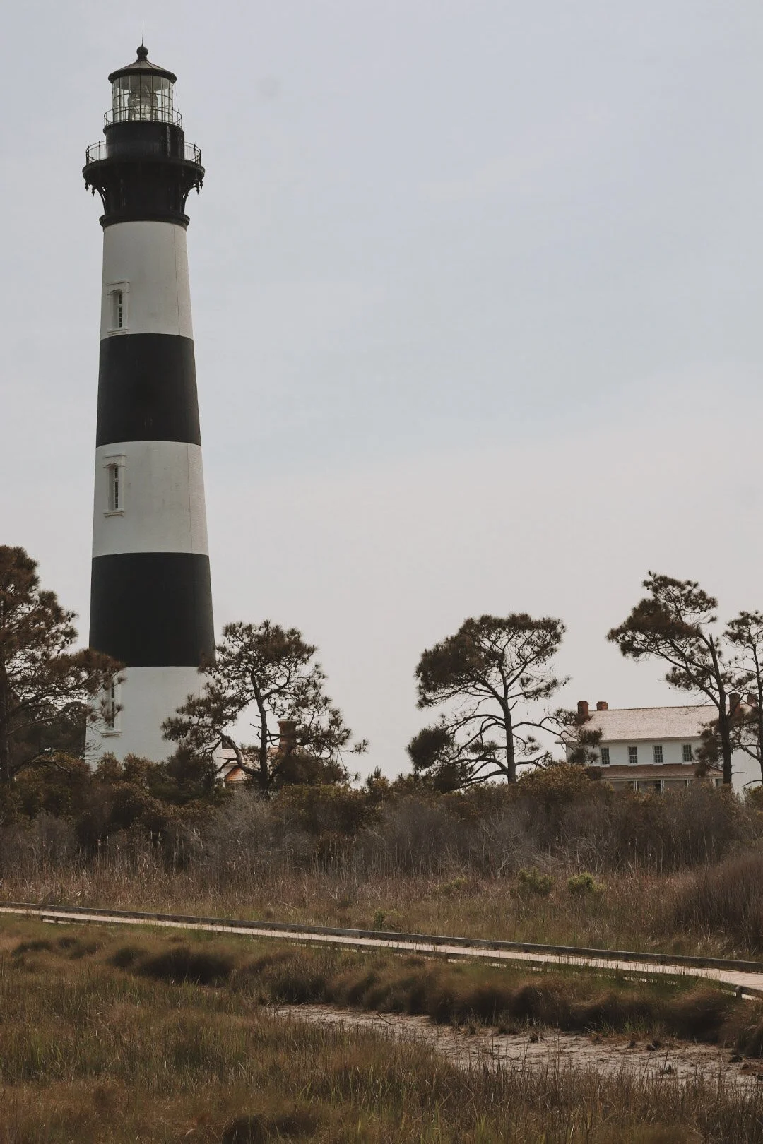 A lighthouse with black and white horizontal stripes, surrounded by trees and a house in the background.
