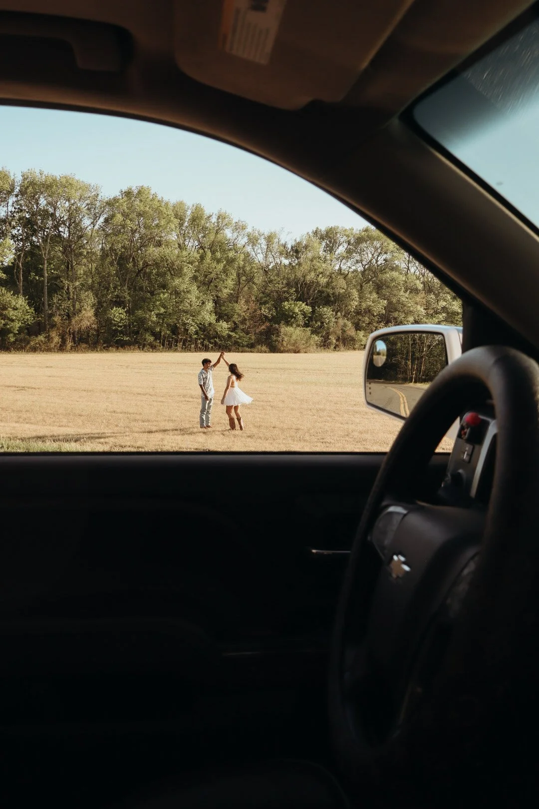 View from inside a vehicle showing two children dancing and playing in a field with trees in the background.