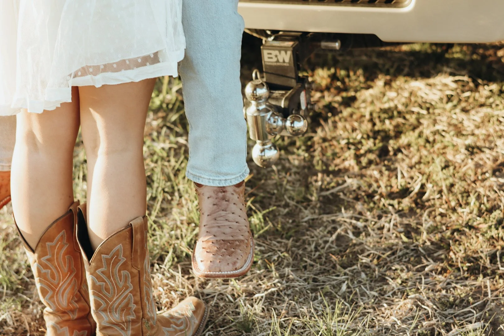 Close-up of two people standing next to a trailer hitch, one wearing cowboy boots and the other in lightly distressed boots, with grass and dirt ground in the background.