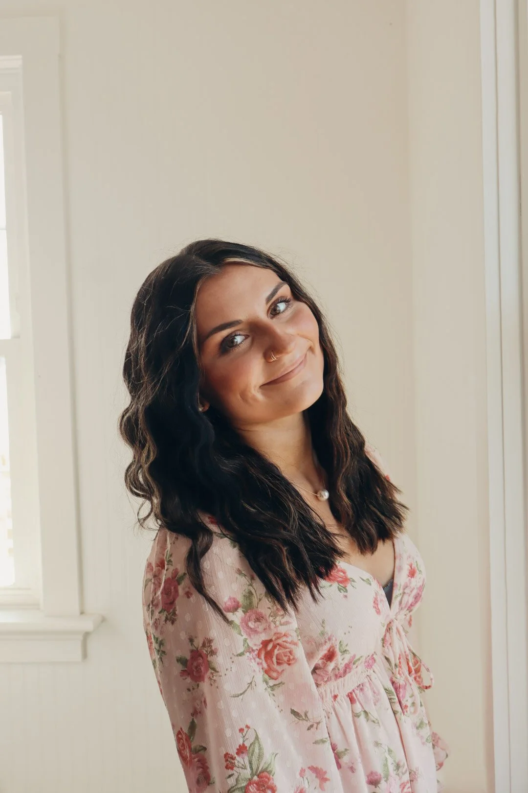 A woman with long, wavy dark hair, light skin, and a nose piercing, smiling slightly, wearing a floral pink dress, standing indoors with light-colored walls and a window.
