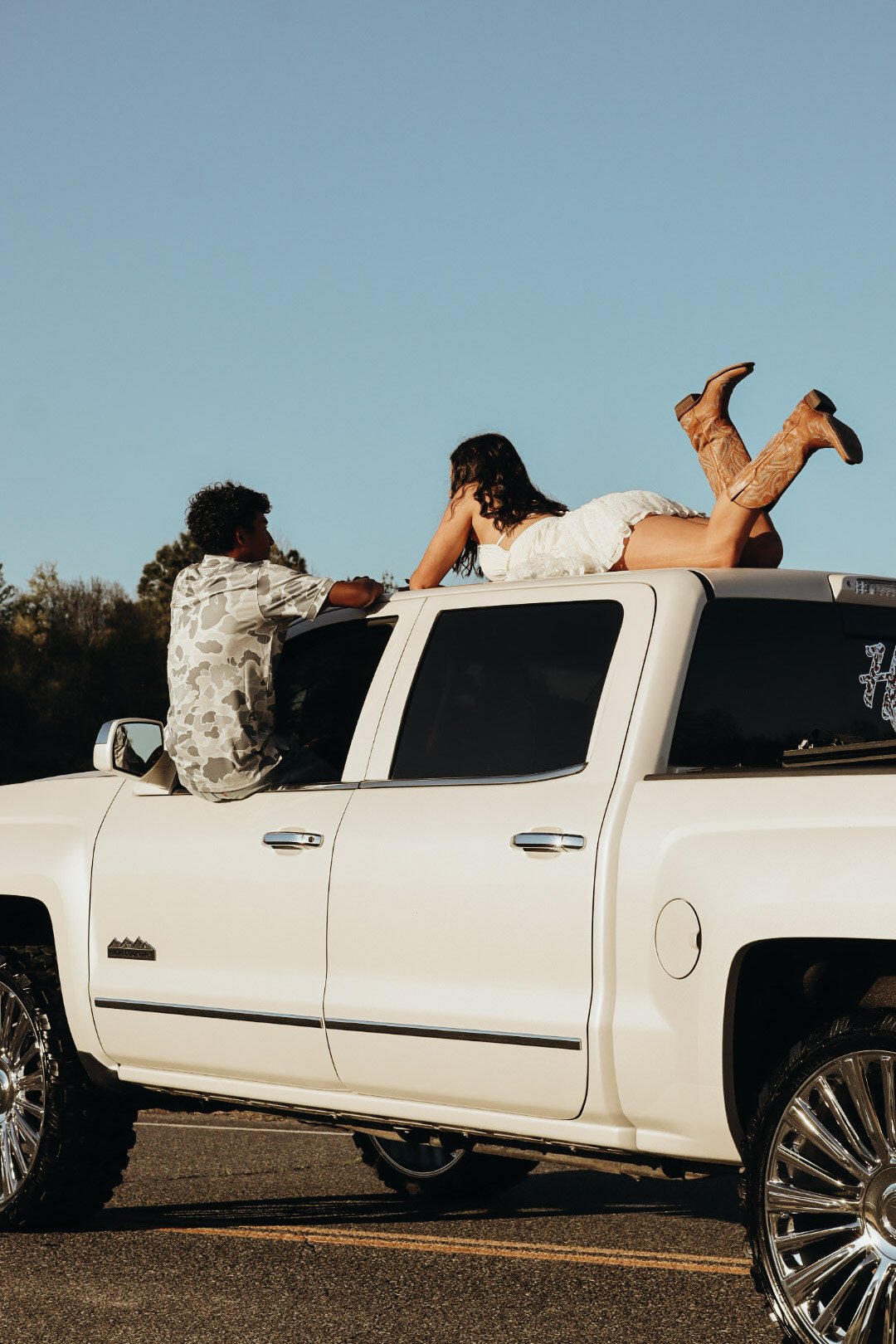 A young woman with dark hair is lying on top of a white pickup truck, wearing a white dress and cowboy boots, while a young boy with curly hair is sitting on the side of the truck, looking at her under a clear blue sky.