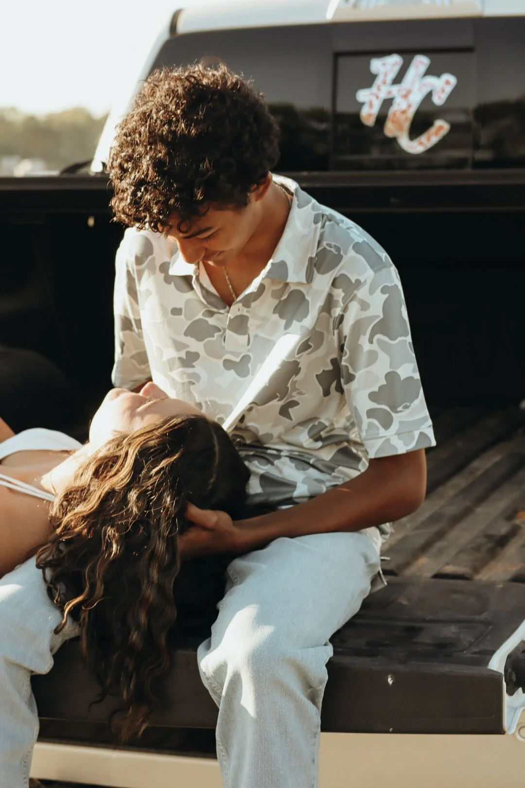 A young man with curly hair wearing a camo shirt holds a woman with long wavy hair in his lap, sitting on the back of a black vehicle with a Glendale, Arizona sign on the rear window. They appear to be happy and affectionate.