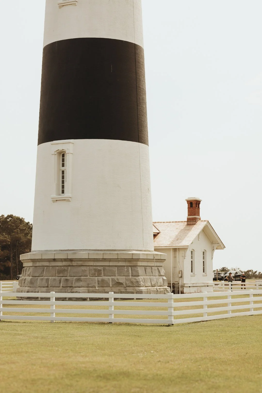 A lighthouse with black and white horizontal stripes next to a small white house with a brick chimney, surrounded by a white picket fence.