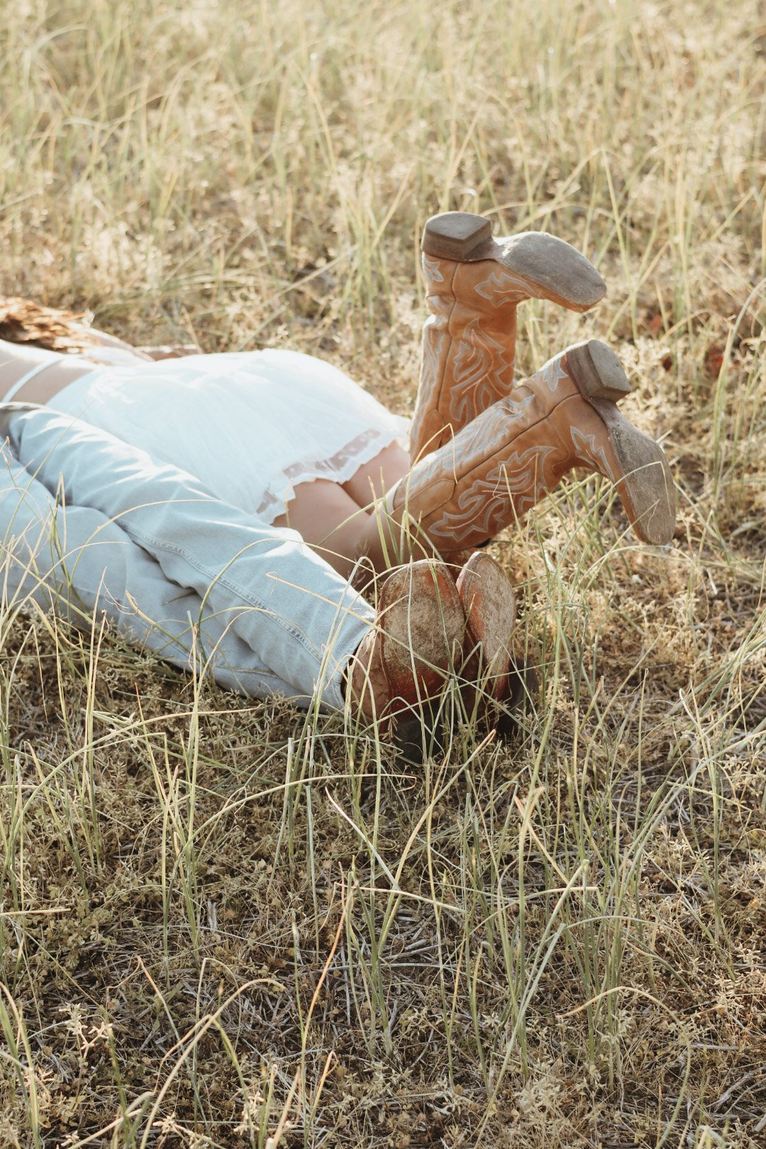 A person lying on their back in a grassy field, wearing jeans, a white shirt, and brown cowboy boots with white decorative stitching.