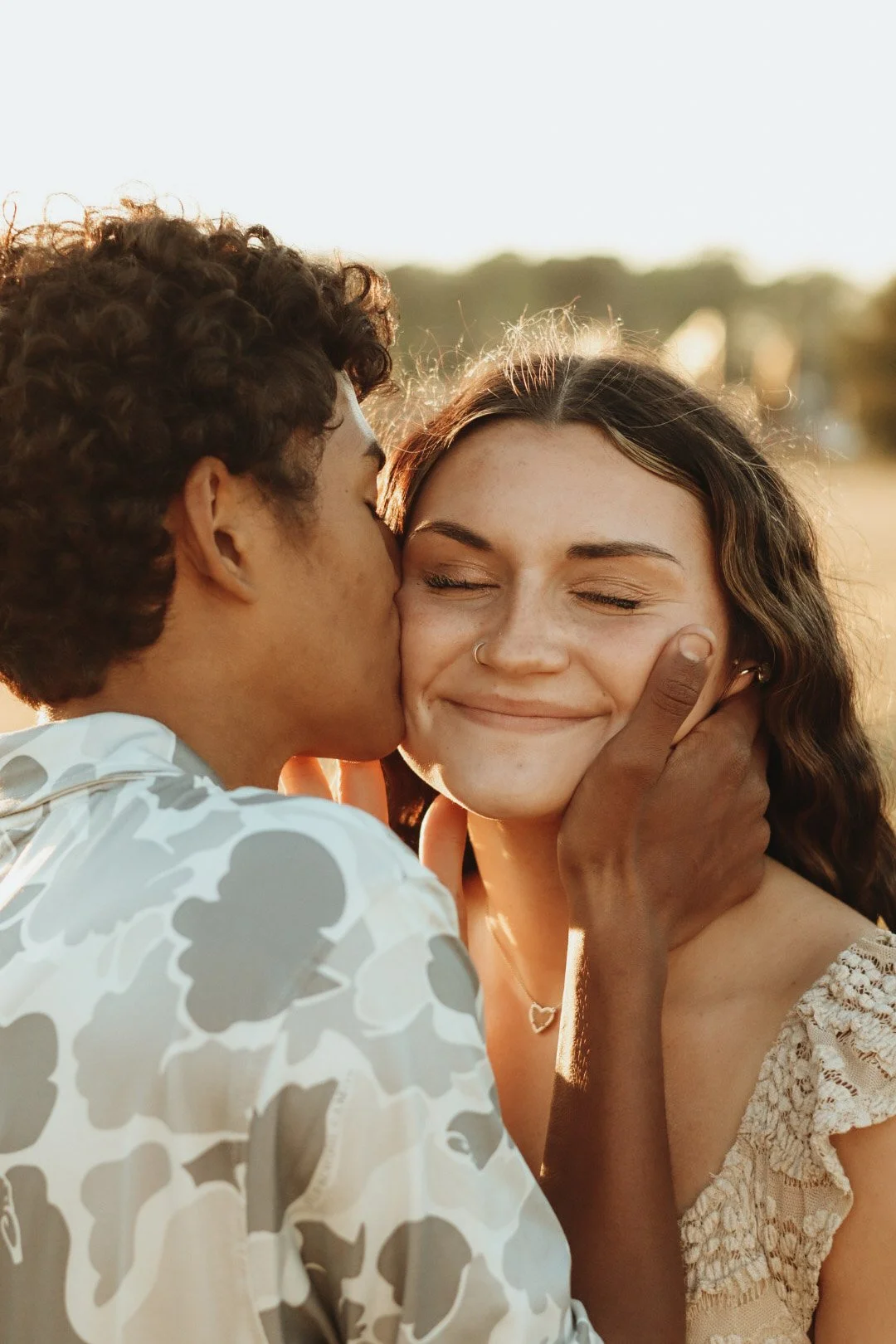 A man kissing a woman on the cheek in a sunset outdoor setting, with the woman smiling and her eyes closed.
