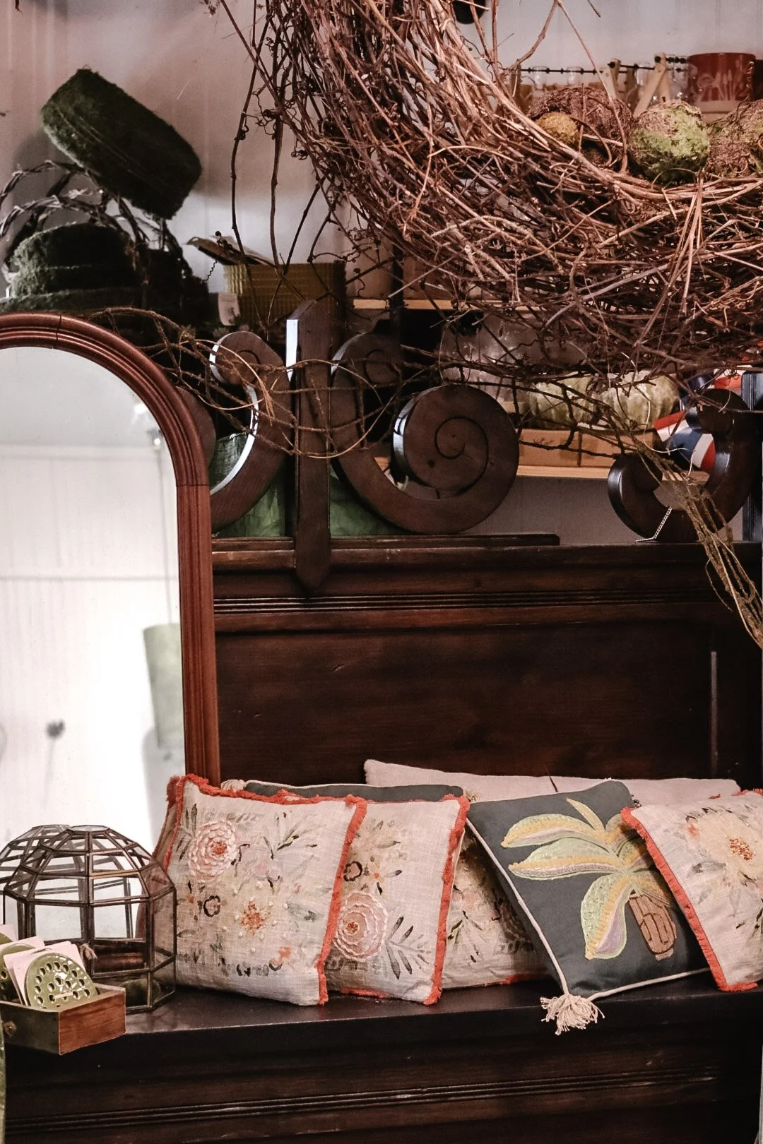 A vintage dresser with floral and leaf-patterned pillows on top, a decorative metal cage, and a large ornate mirror on the left. A large, messy bundle of twigs or branches rests on the dresser, with some other decorative items in the background.