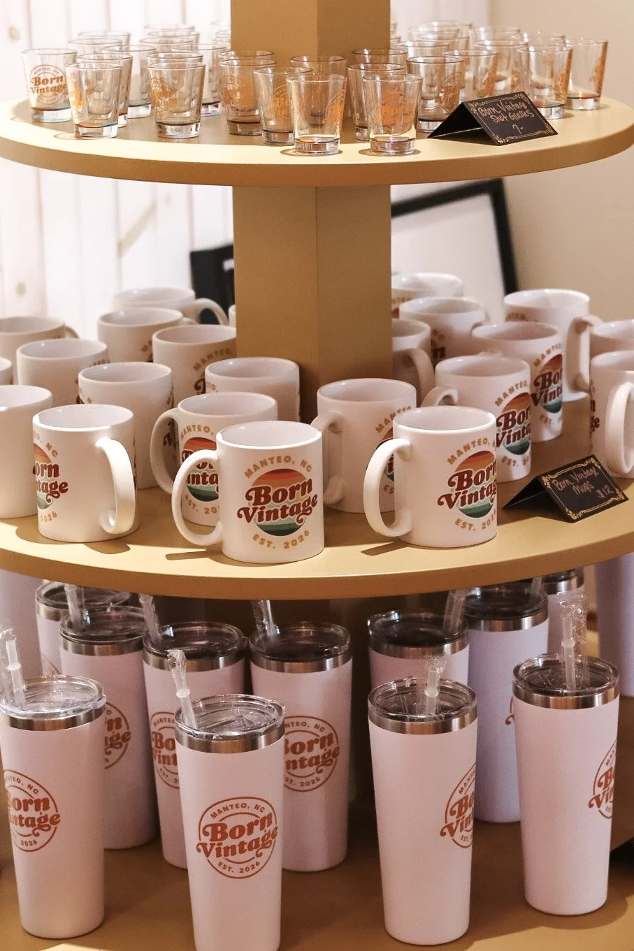 Display of white mugs and tumblers with 'Born Vintage' branding on a yellow, three-tiered stand at a promotional event.