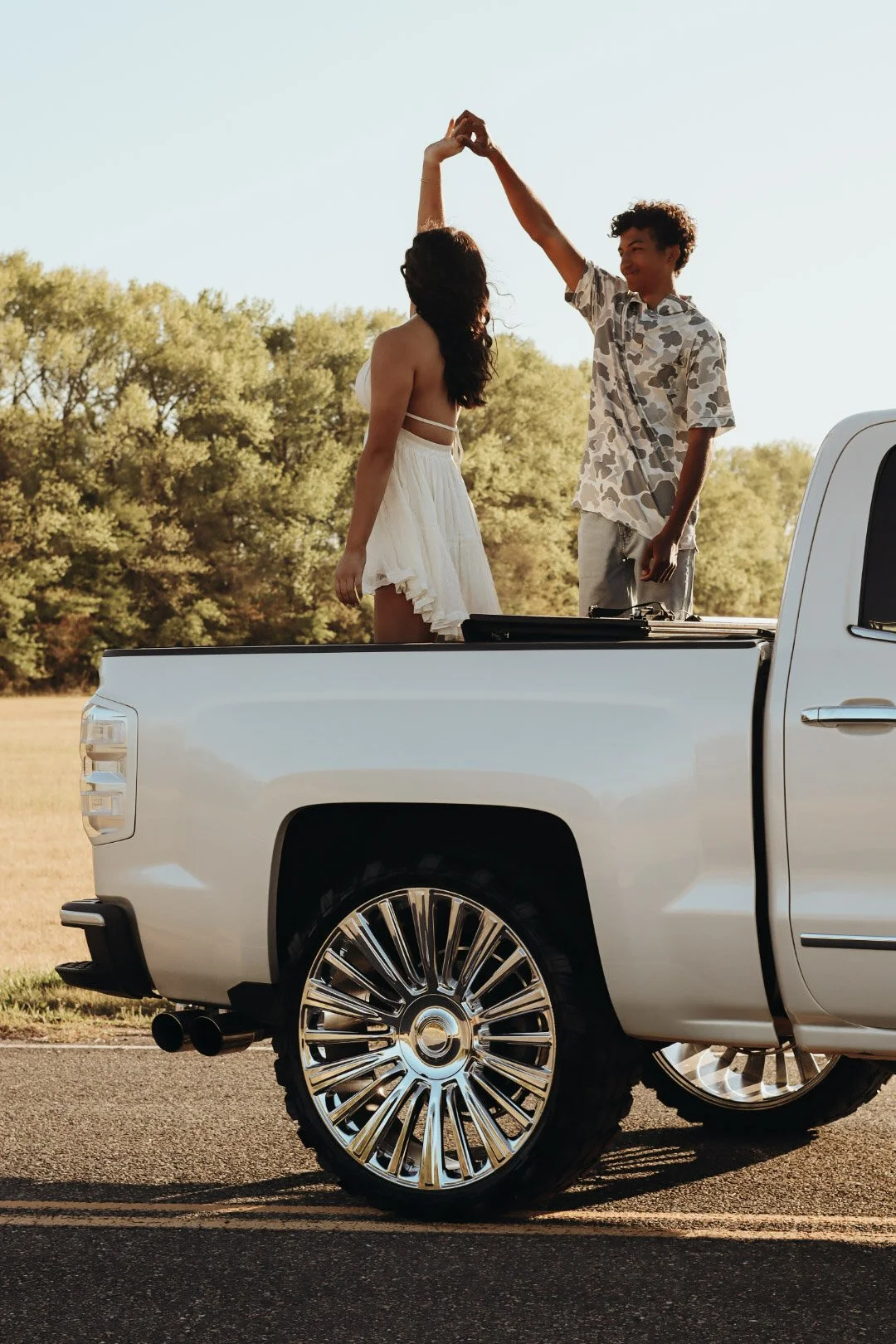 A young couple dancing in the back of a white pickup truck outdoors on a sunny day, with trees in the background.