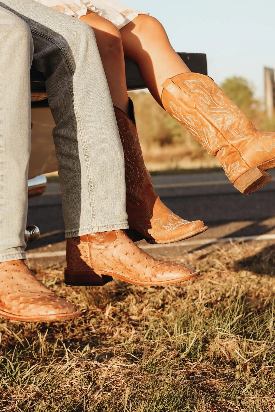 Two people sitting on a bench outdoors, both wearing cowboy boots and denim jeans, with their legs crossed or dangling over the edge of the bench, on a grassy and dirt area with trees and a road in the background, during sunset.