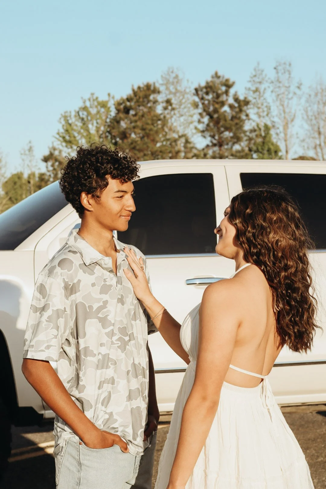 A young man and woman facing each other outdoors near a white vehicle, with the woman touching the man's chest, both smiling as they look at each other, with trees in the background.