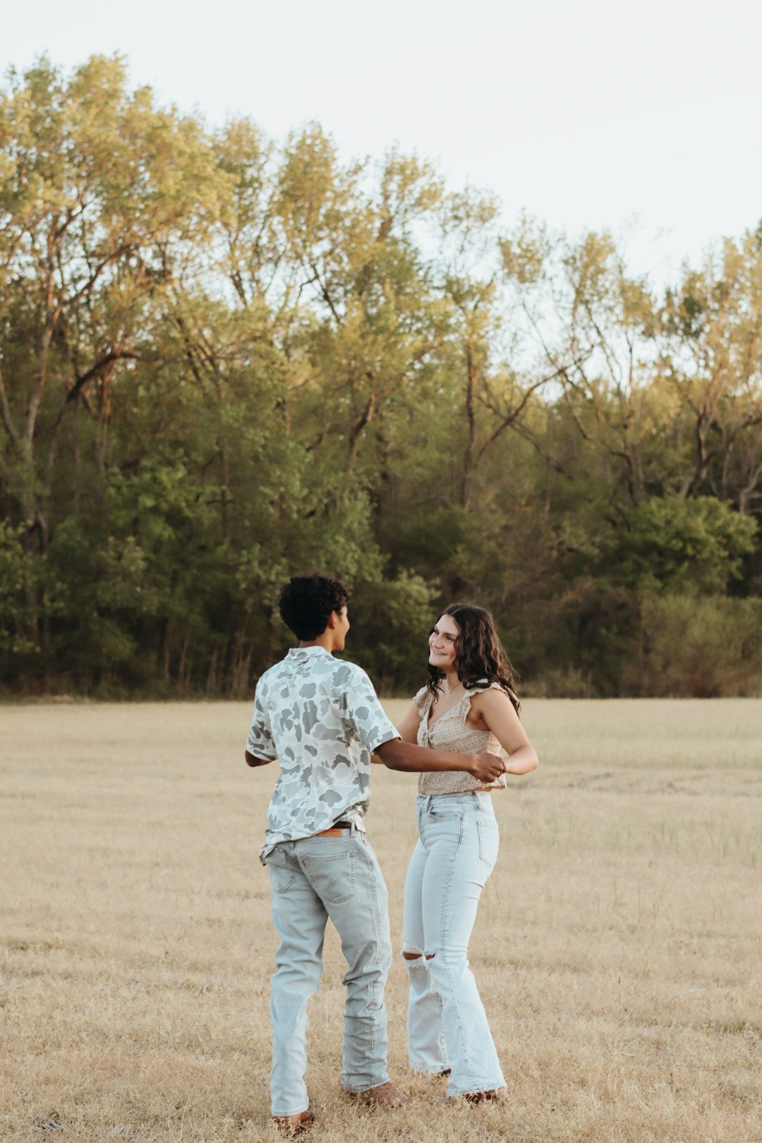 A young couple is dancing outdoors in a field, holding hands and looking at each other, with a background of trees and a clear sky.