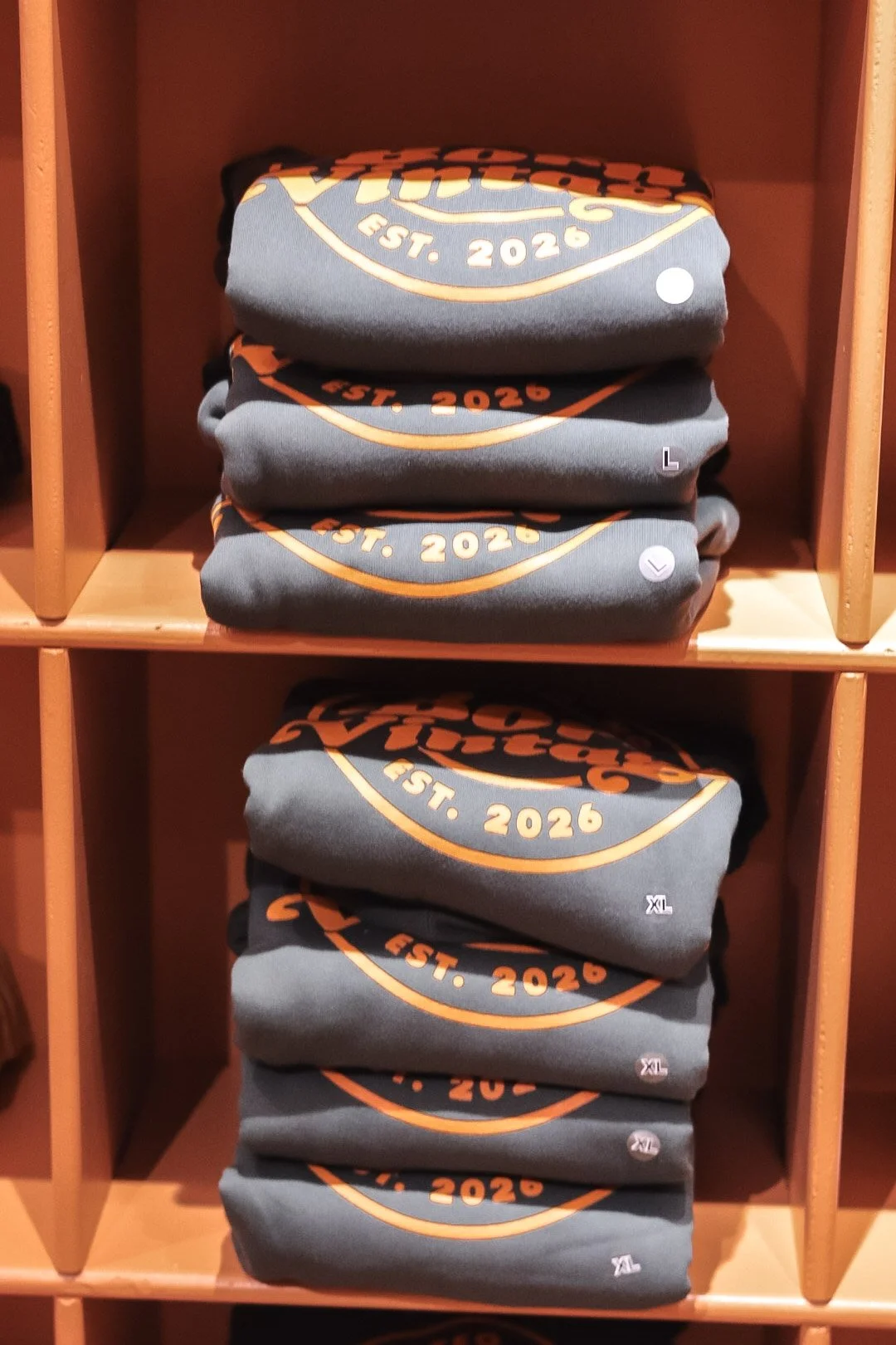 Folded grey sweatshirts with orange and white design and text reading "EST. 2026" on a wooden shelf, with size labels XL, L, and T4 visible.