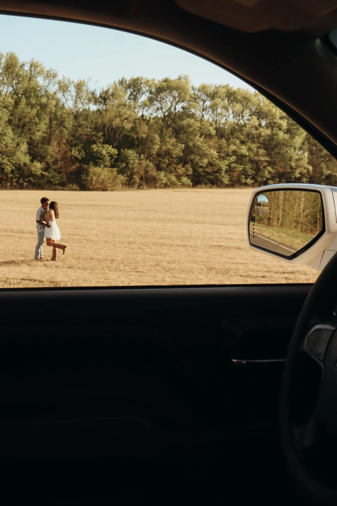 A couple standing in a field holding hands, viewed from inside a car with the side mirror visible.