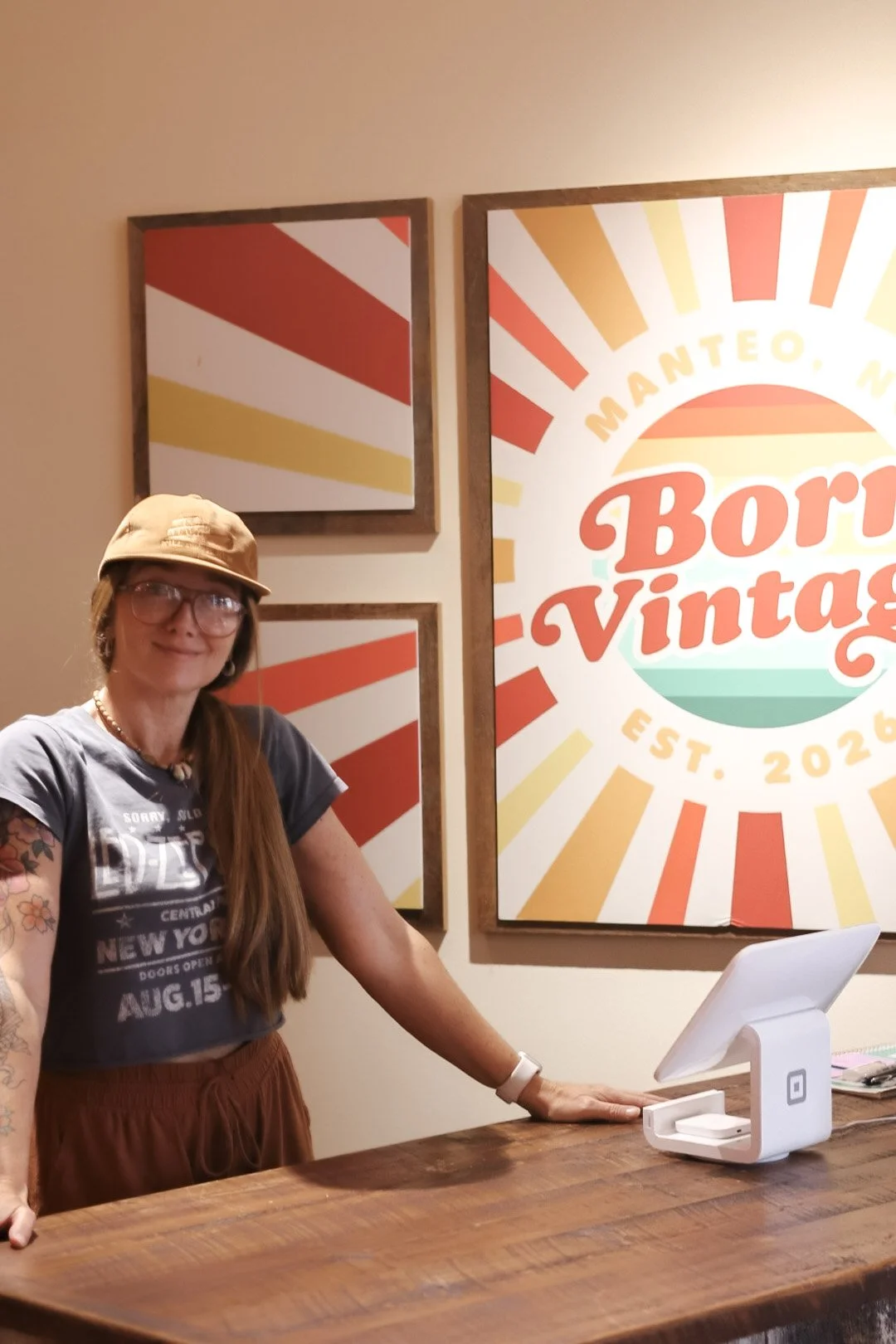 A woman with long hair, glasses, a tan cap, and tattoos on her arm, standing at a wooden counter with a white point-of-sale device, inside a store with vintage-themed decor.