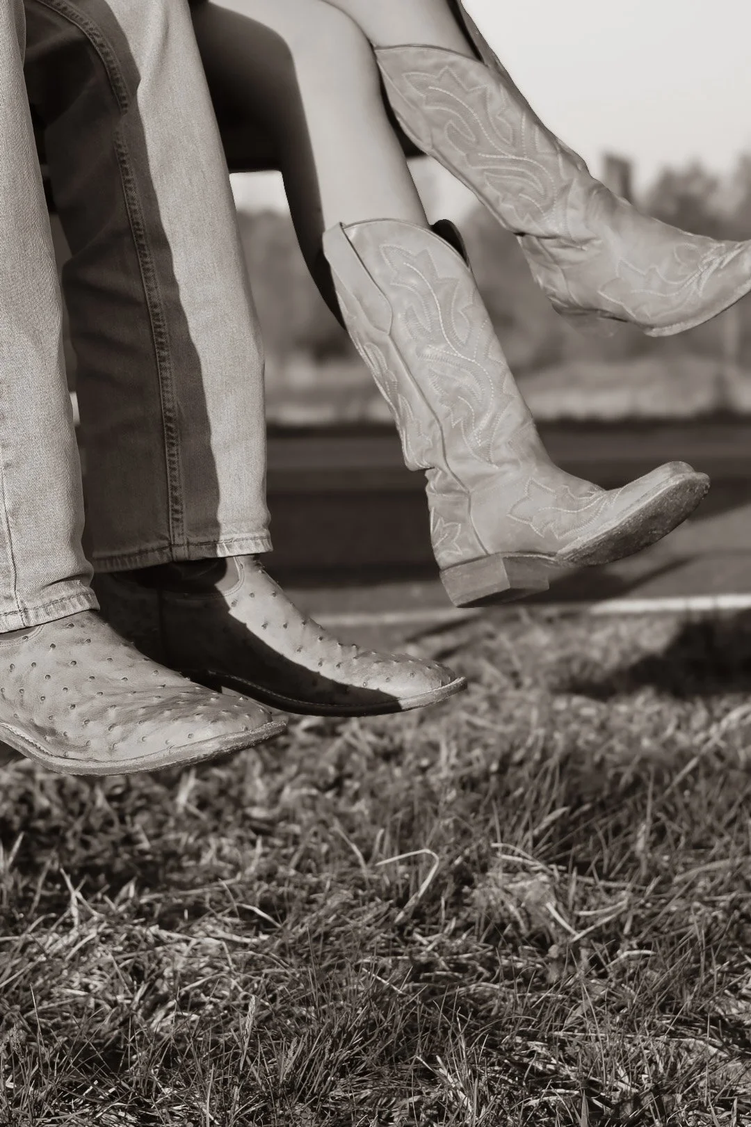 Close-up of two cowboy boots and jeans, one person is sitting while the other is standing, with grass and a blurred landscape in the background, black-and-white photo.