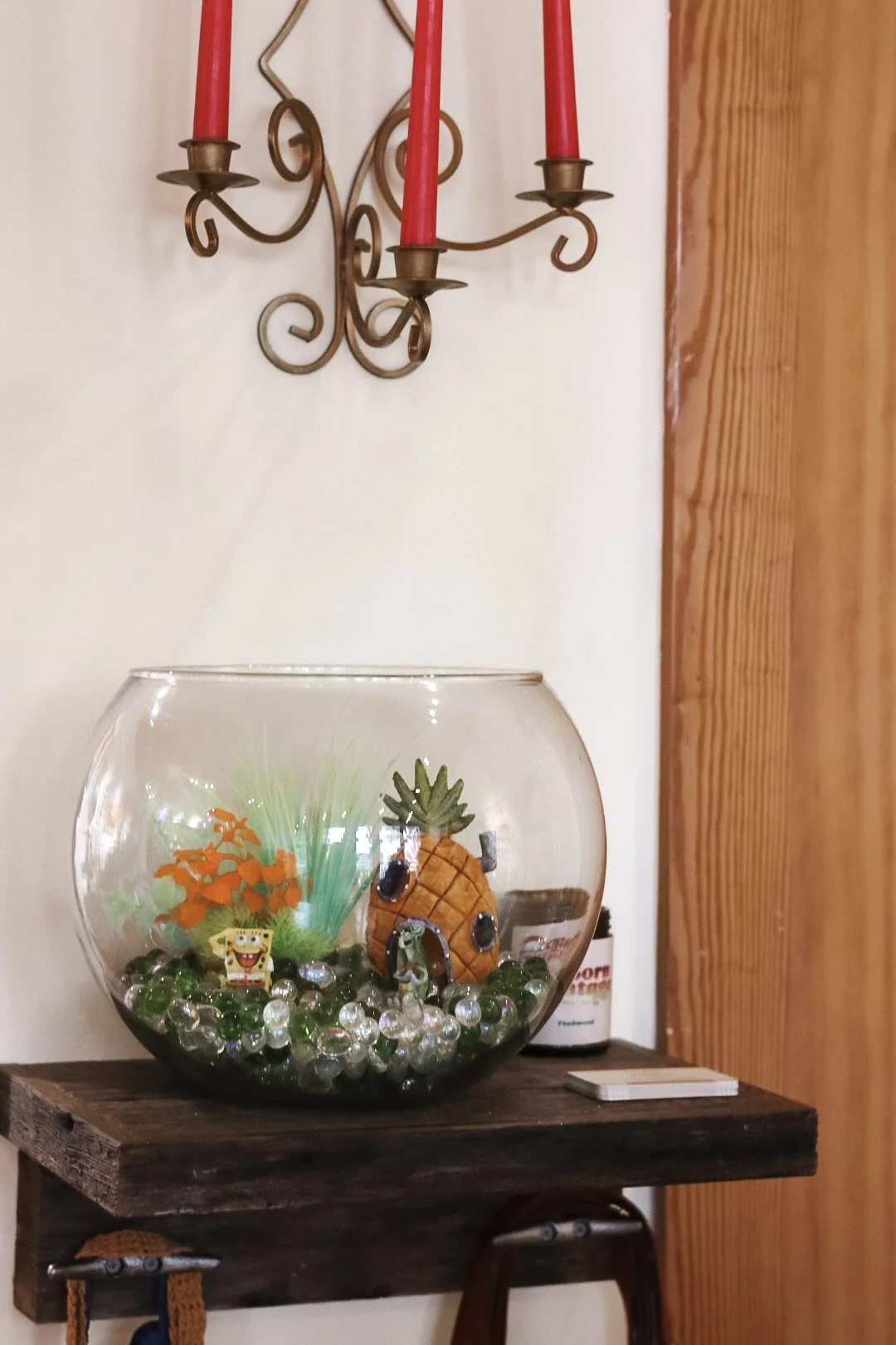 Decorative glass bowl with a pineapple-shaped ornament, orange plastic plants, and green marbles, placed on a wooden table below a wall-mounted iron candle sconce with red candles.