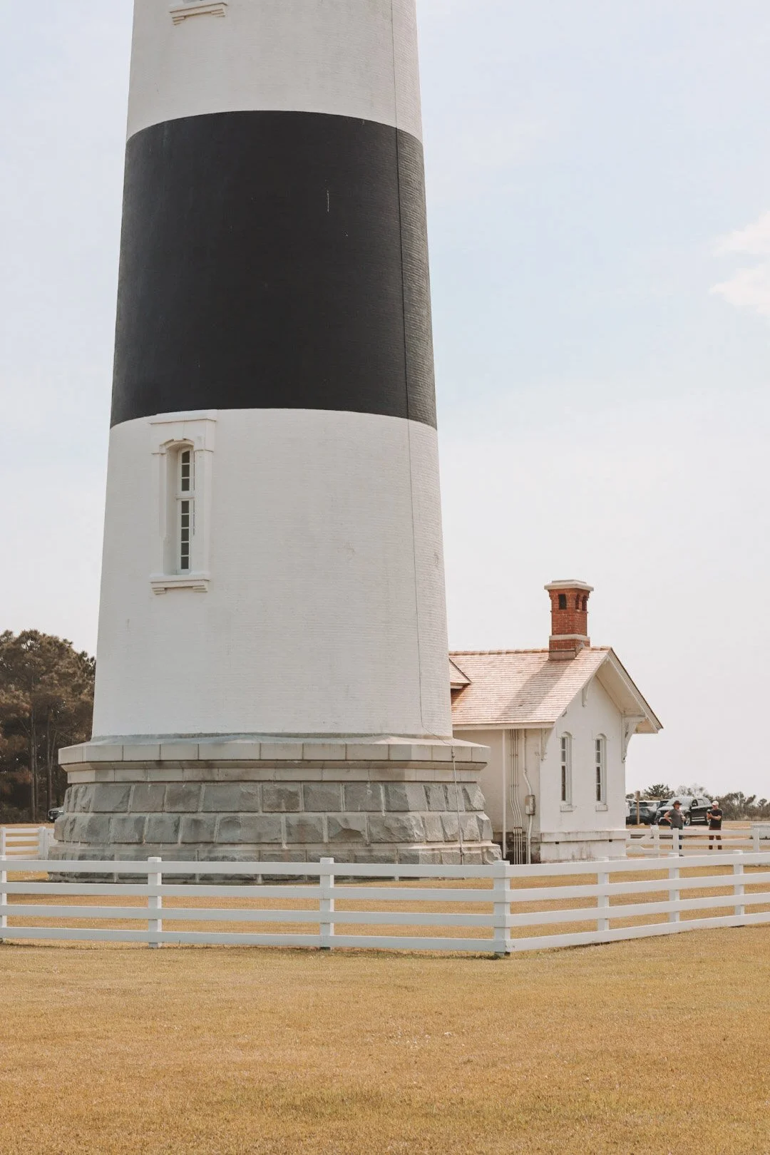 Lighthouse with black and white horizontal stripes, brick house with chimney nearby, surrounded by a white picket fence, grassy area, and people in the background under a partly cloudy sky.