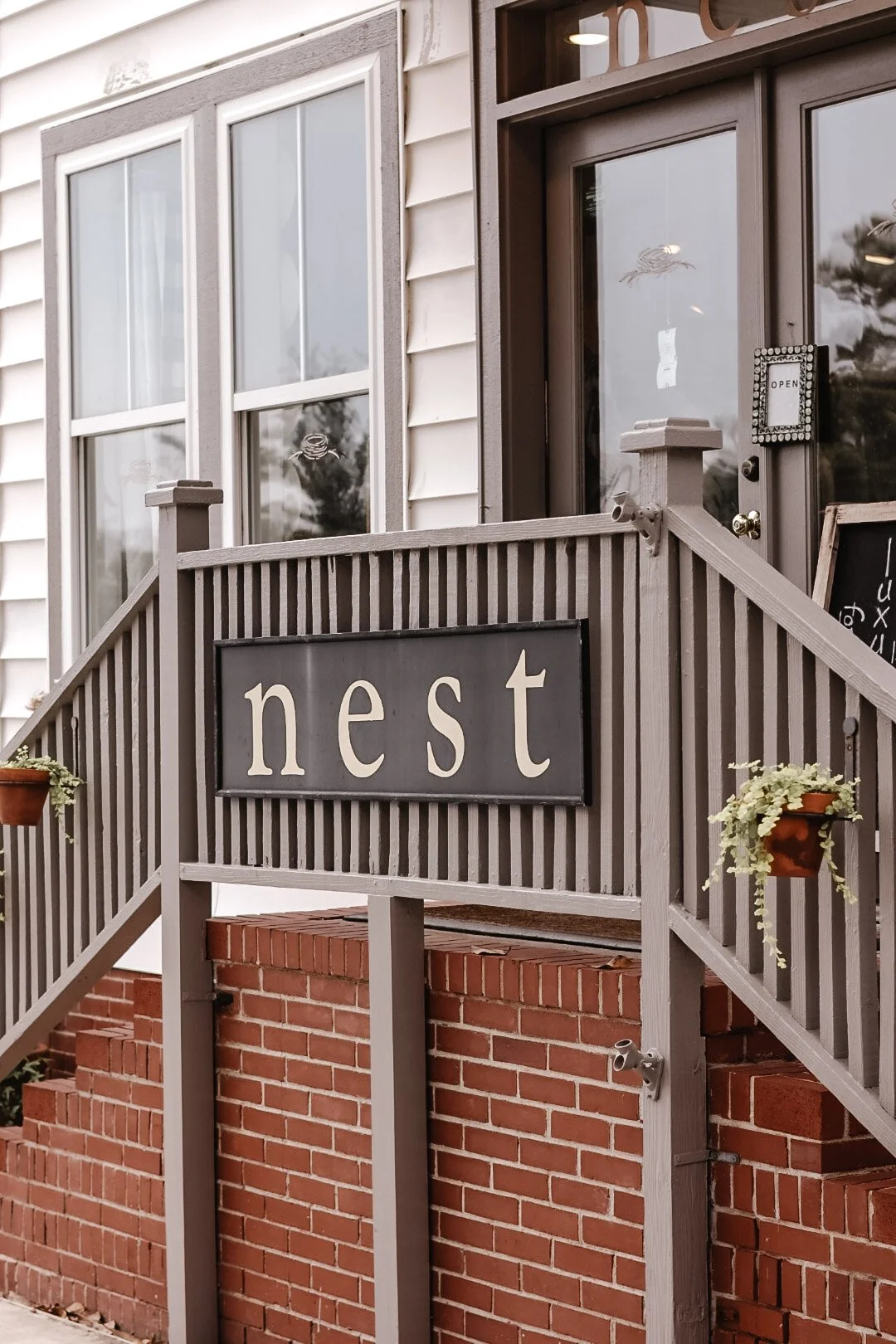A storefront with a sign that says 'nest'. The entrance has stairs, a railing, and a door with a small sign that says 'open'. There are two windows and some small hanging flower pots.
