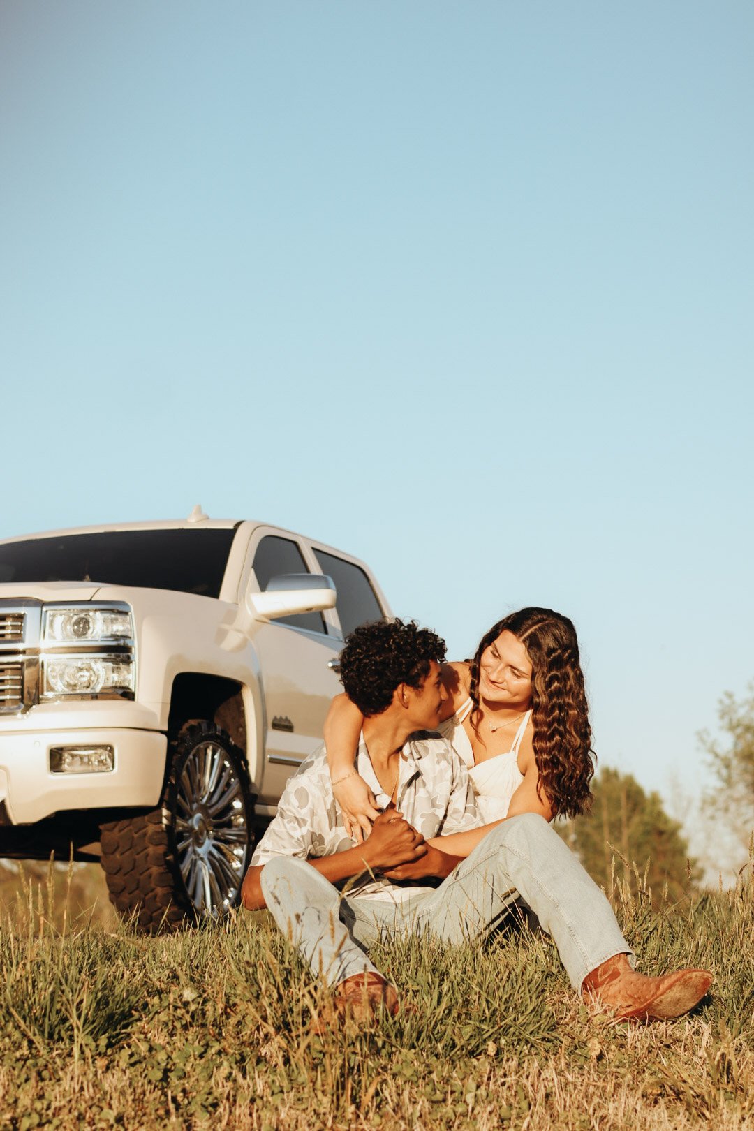 A couple sitting on the grass, embracing and looking into each other's eyes, next to a white luxury SUV, under a clear blue sky.