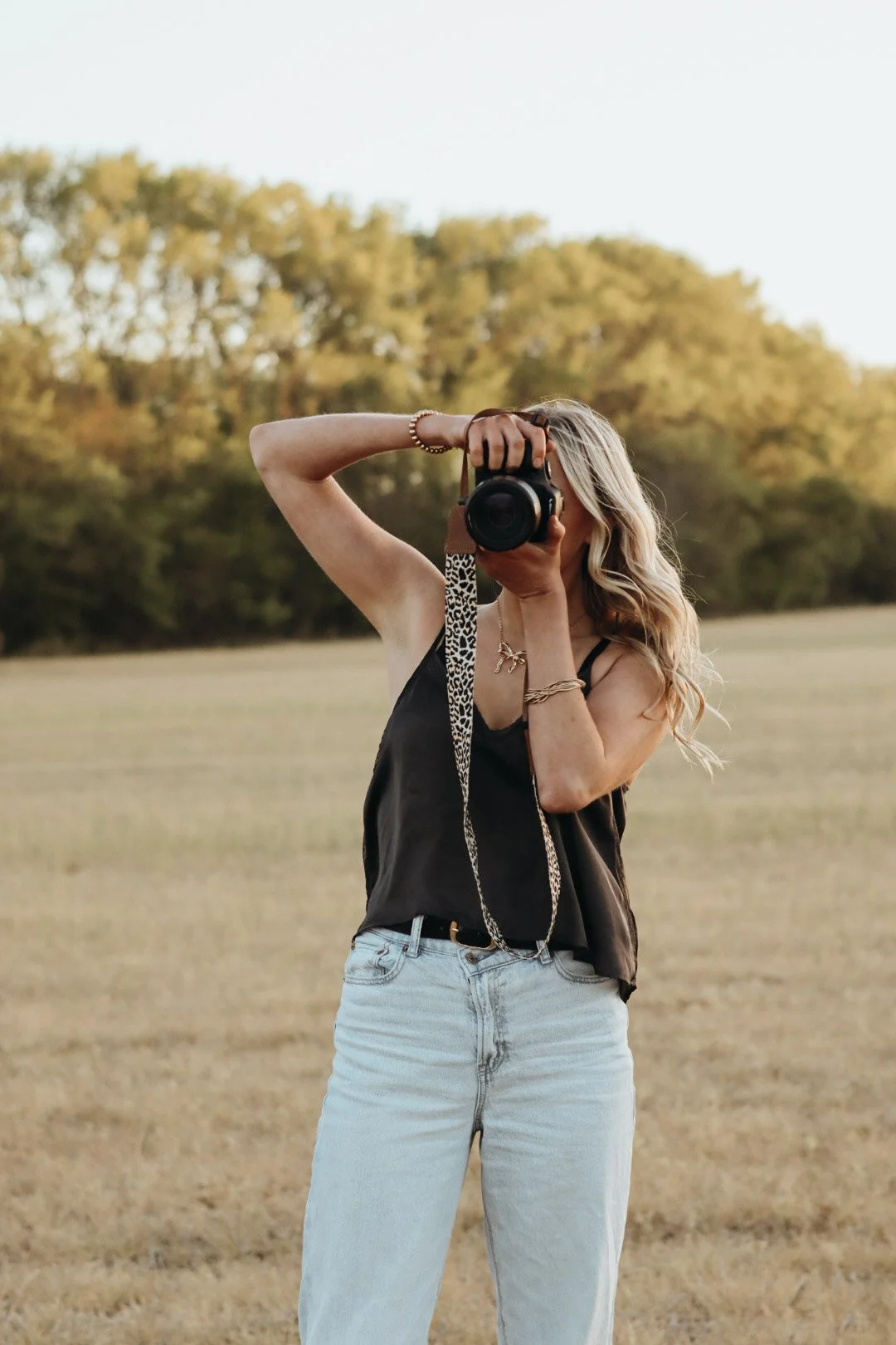 A woman with long blonde hair taking a photo outdoors with a camera, standing in a field with trees in the background during the daytime.