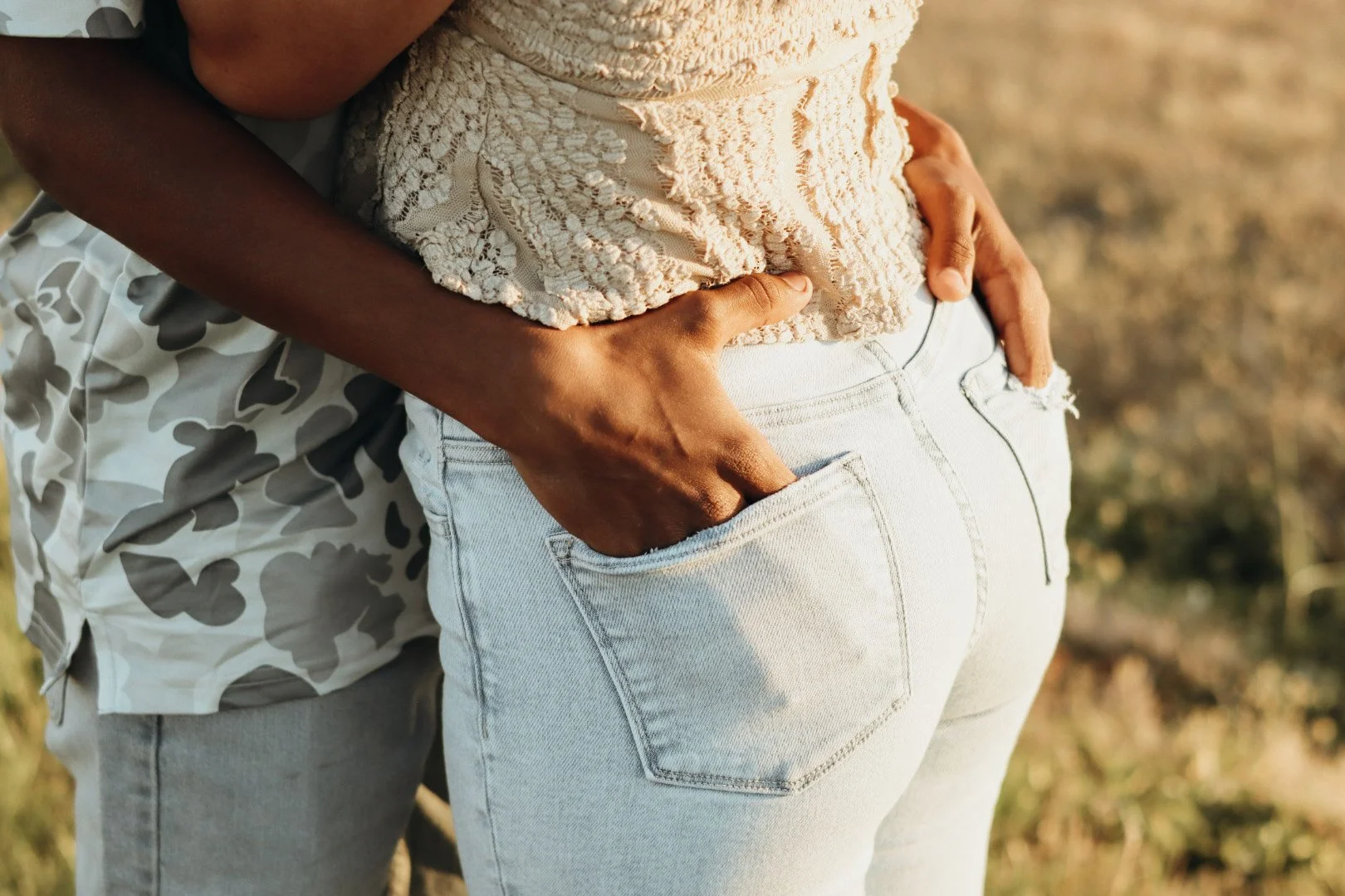 Close-up of a person's hand resting on the back pocket of another person's white jeans, with the person wearing a beige lace top or cover-up. They are outdoors in a natural setting.