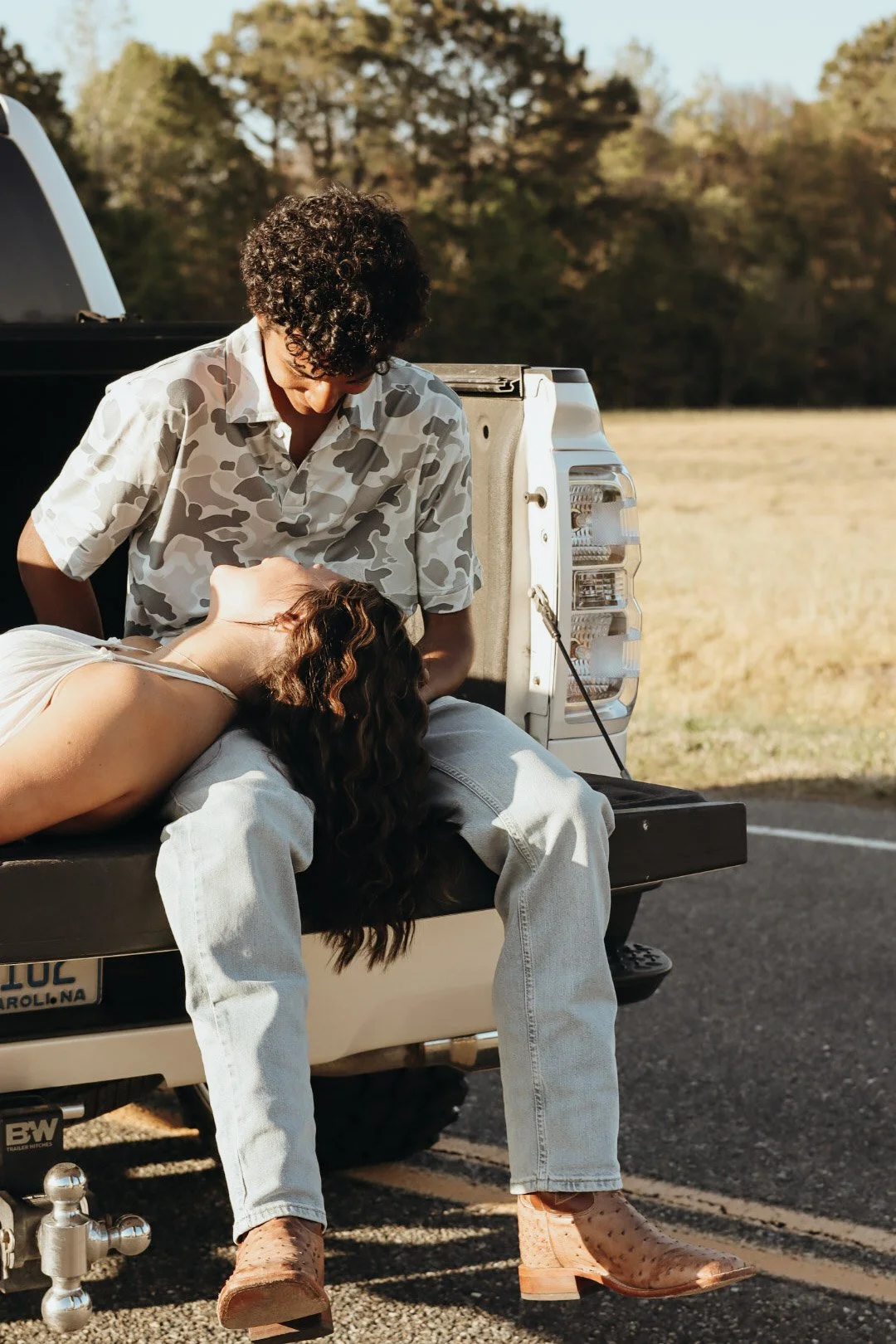 A young man and woman are sitting on the tailgate of a pickup truck in a rural, outdoor setting during golden hour. The man has curly hair and is wearing a camouflage shirt, leaning over the woman who is lying back with her head on his lap. The woman
