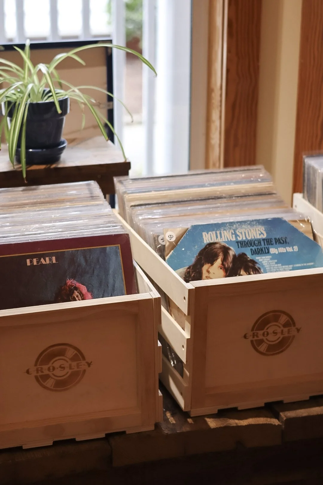 Wooden crates filled with vinyl records, including albums by The Rolling Stones and Pearl, on a wooden table beside a potted plant, indoors near a window.
