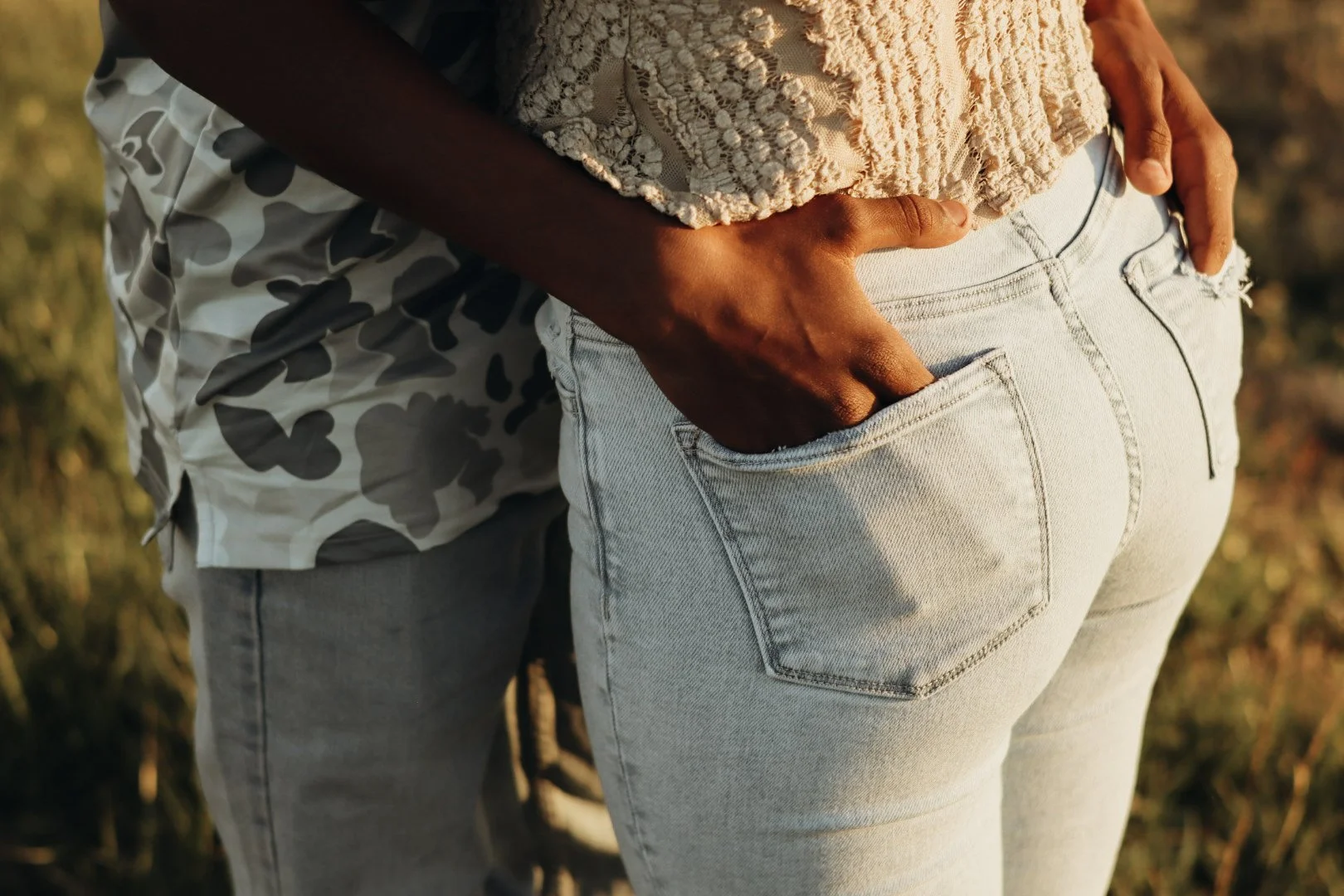 Close-up of two people standing closely, one with hand in the back pocket of light-colored jeans, outdoor setting with warm sunlight.