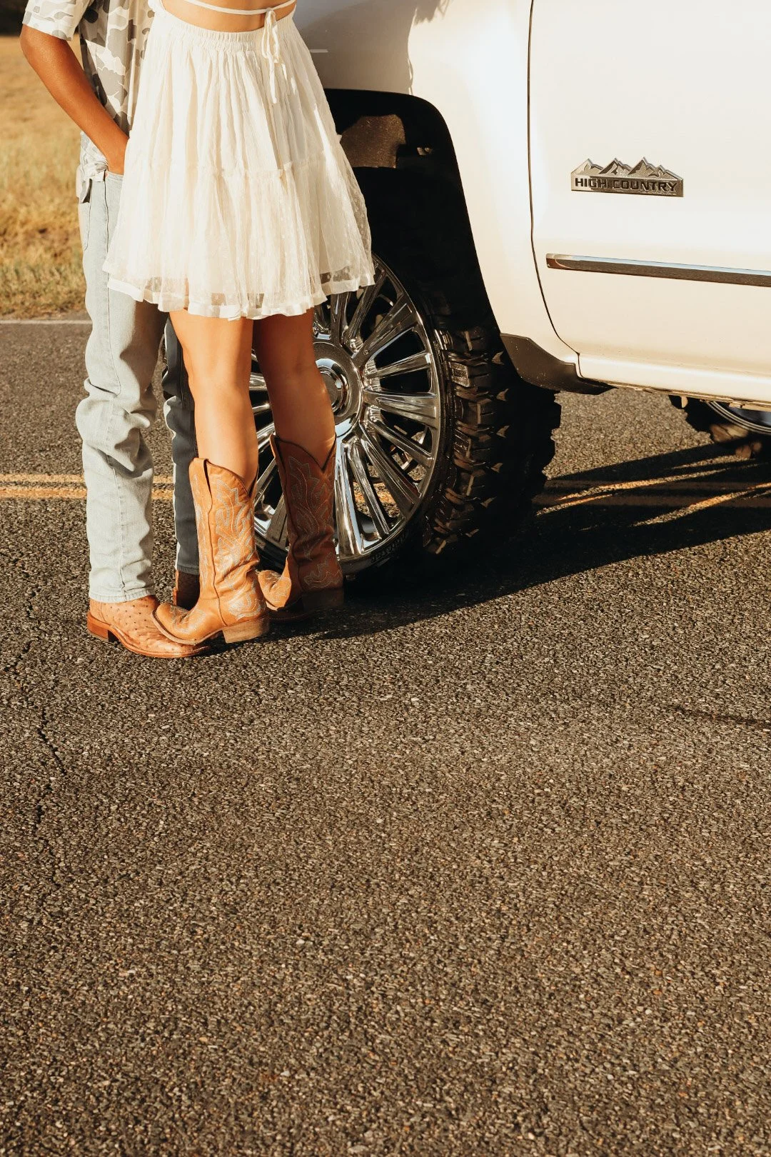 Close-up of a woman wearing a white skirt and cowboy boots standing next to a white high country pickup truck. Only part of a man is visible, standing behind her with light-colored jeans. The photo is taken on a paved road during sunset.